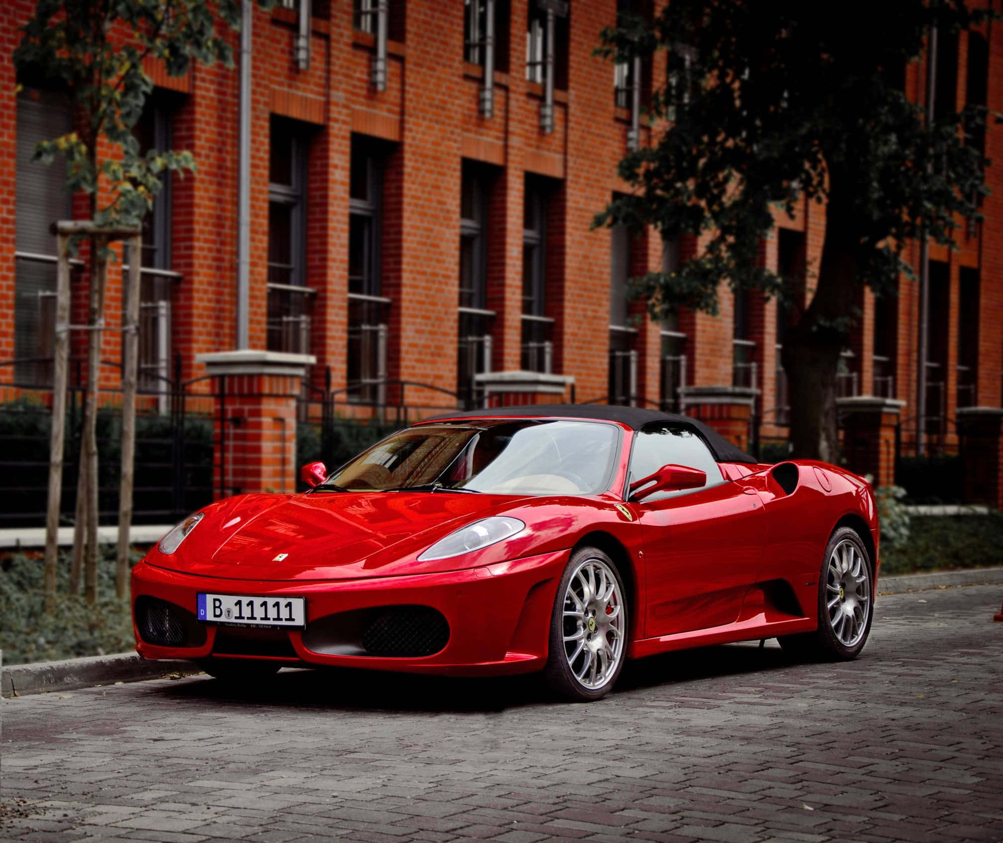 Ferrari 360 Modena 桌布 壁紙 — 一輛光亮的紅色法拉利Spider停泊在鵝卵石街道上，前面是帶樹木的磚砌建築。