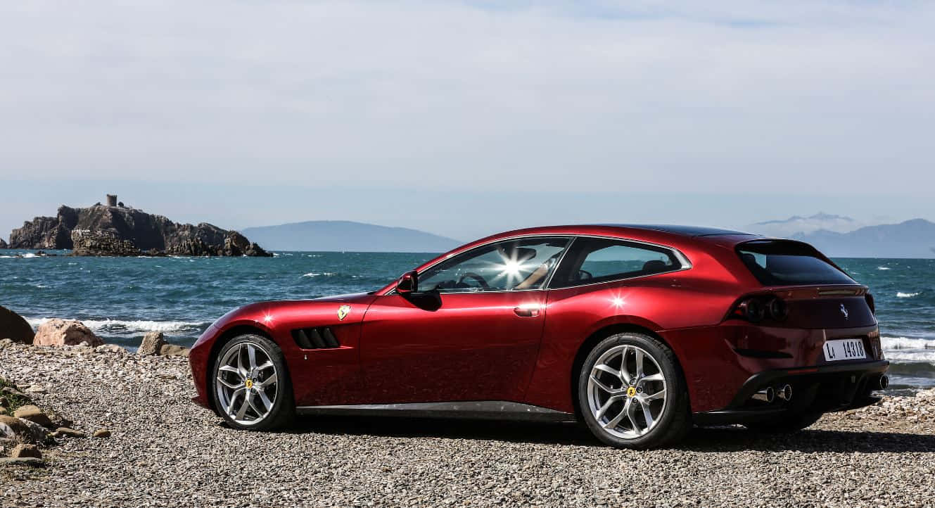 Ferrari Gtc4lusso 桌布 壁紙 — 一輛光亮的紅色法拉利跑車停泊在岩石海灘上，海浪拍打，遠處懸崖在明亮的天空下。