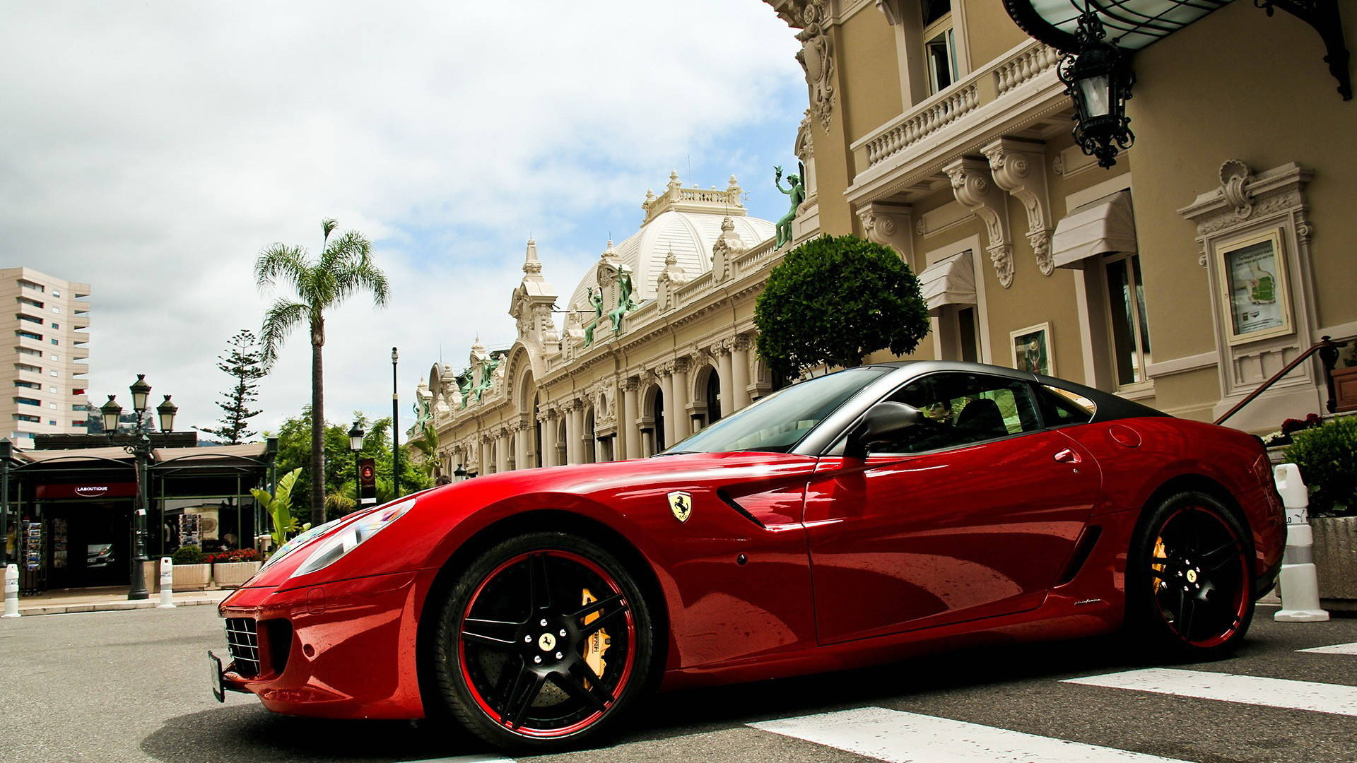 1920x1080 Ferrari 桌布 壁紙 — 一輛光亮的紅色法拉利跑車停泊在街上，旁邊是裝飾華麗的米色建築，天空多雲。