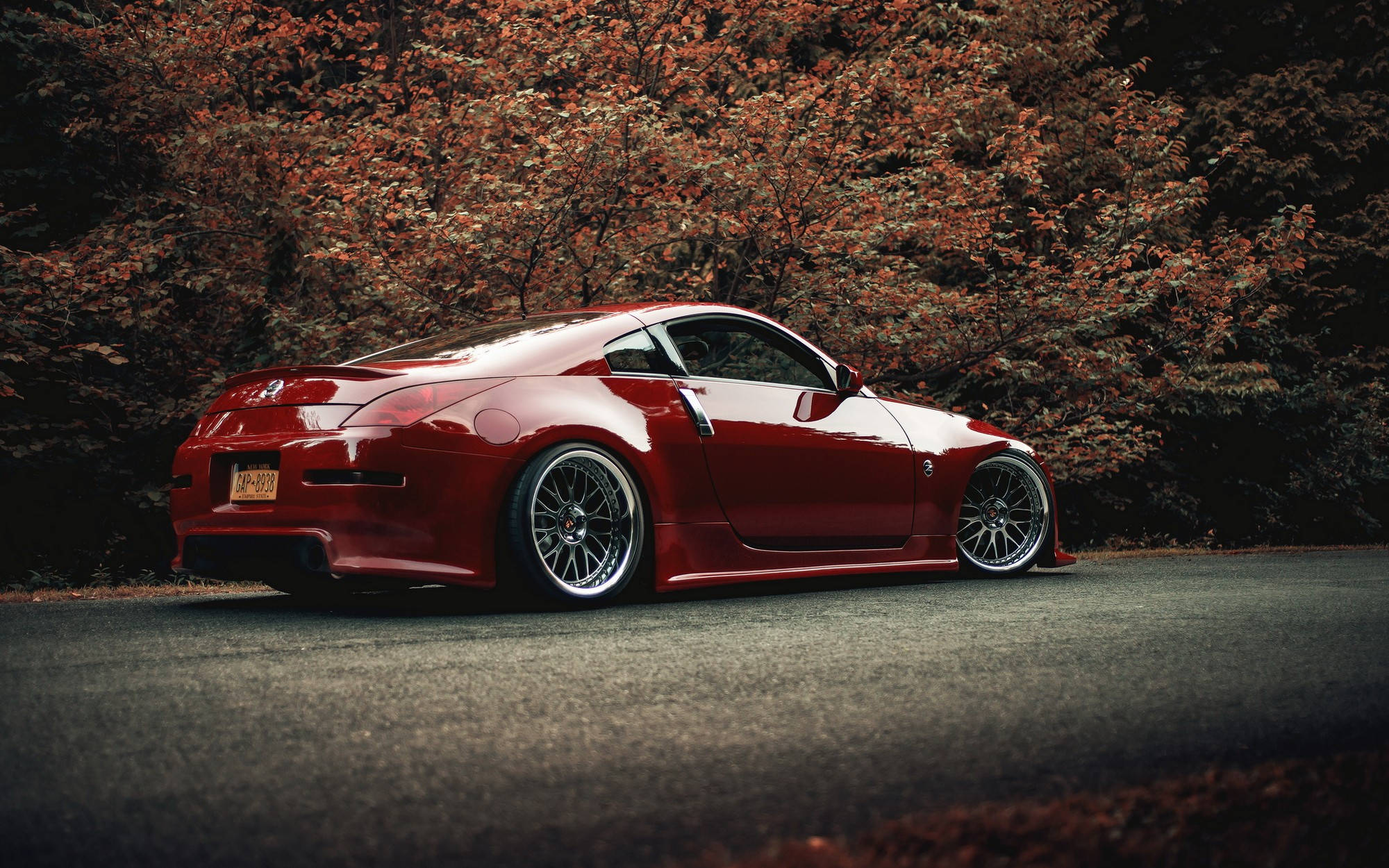 Nissan 350z 桌布 壁紙 — 一輛光亮的紅色跑車配備定制輪圈，停在秋季樹葉覆蓋的瀝青路面上。