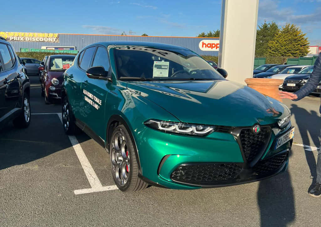 愛快羅密歐 桌布 壁紙 — 一輛光亮的綠色Alfa Romeo Tonale SUV停放在經銷商場地，天空晴朗湛藍，附近有人正在做手勢。