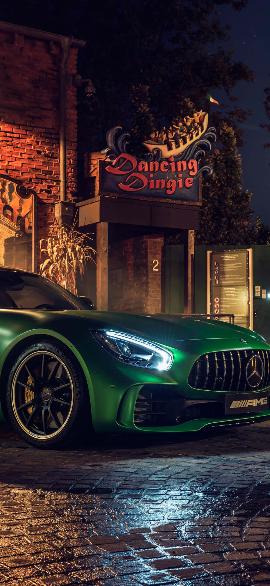 iPhone X Amg Gt-r 桌布 壁紙 — 一輛光亮的綠色AMG跑車在霓虹燈下閃耀，停在夜間濕漉漉的鵝卵石街道上，靠近一棟帶有 'Dancing Dingie' 標誌的磚砌建築。