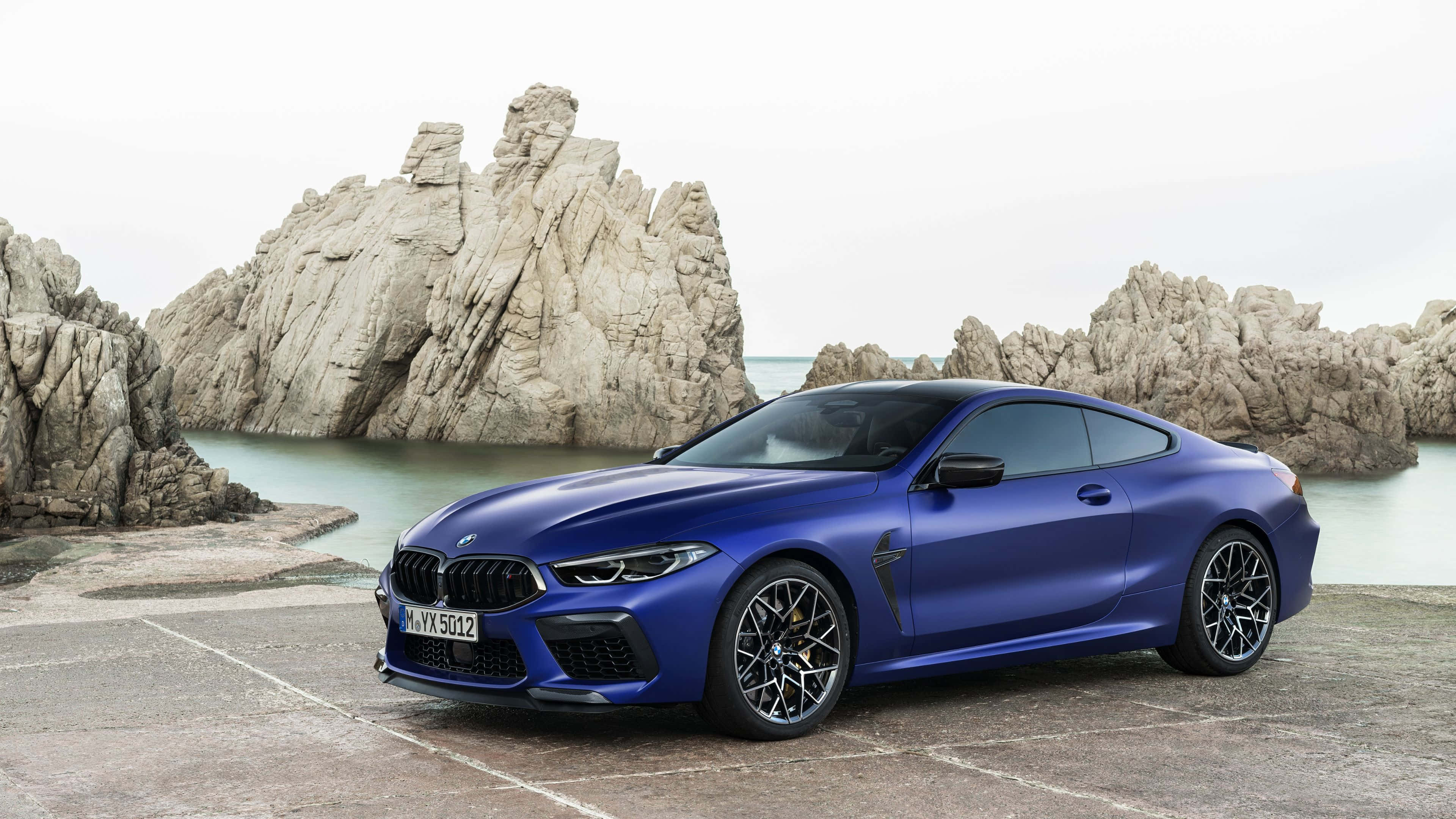 4K BMW 壁紙 桌布 壁紙 — 一輛光亮的藍色BMW M8 Coupe停泊在石碼頭上，旁邊是崎嶇的海岸懸崖和風平浪靜的大海，天空多雲。