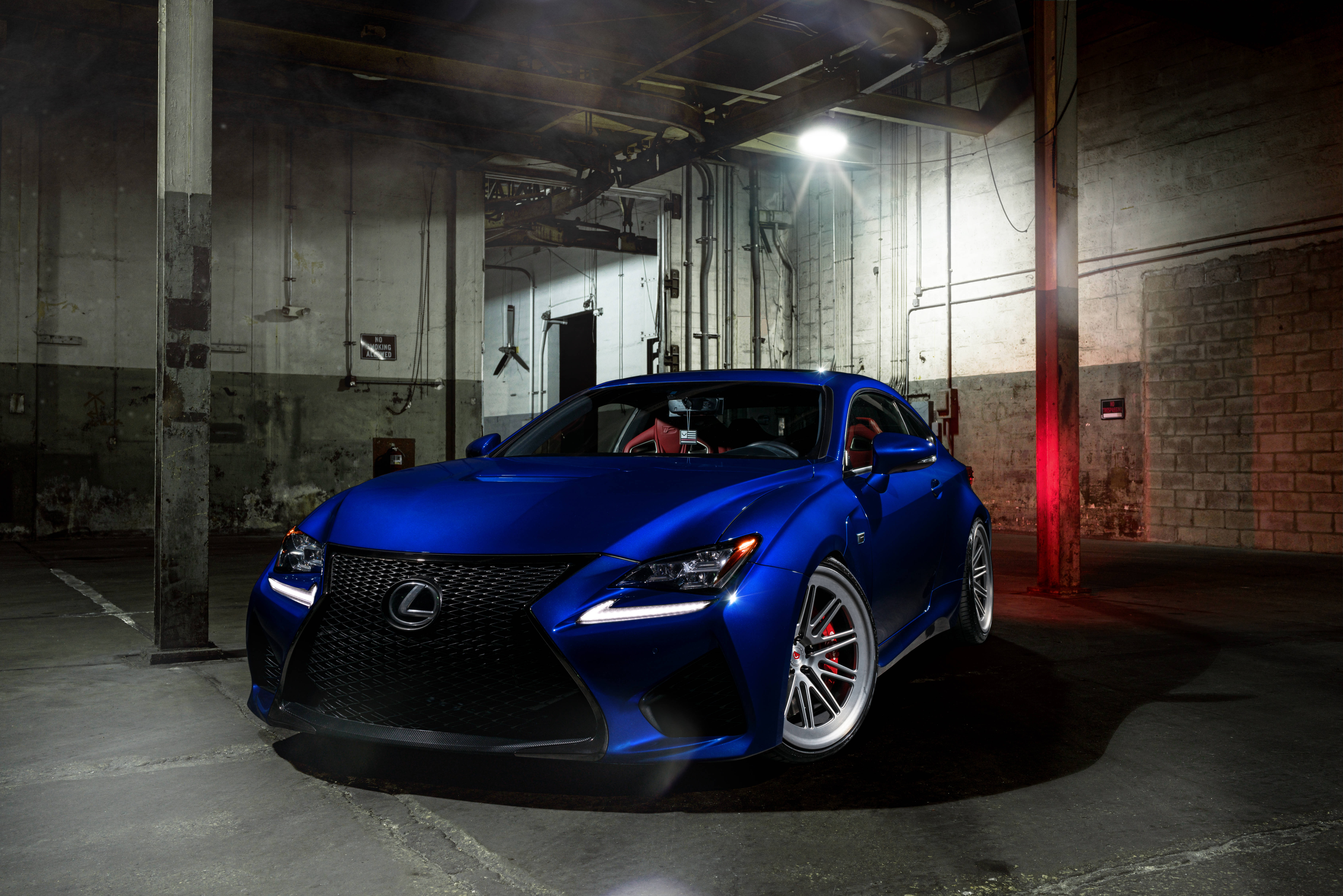 lexus iPhone 桌布 壁紙 — 一輛光亮的藍色Lexus跑車在一個粗糙的工業車庫中，在單一聚光燈下閃閃發光，車庫內有外露的管道和混凝土牆。