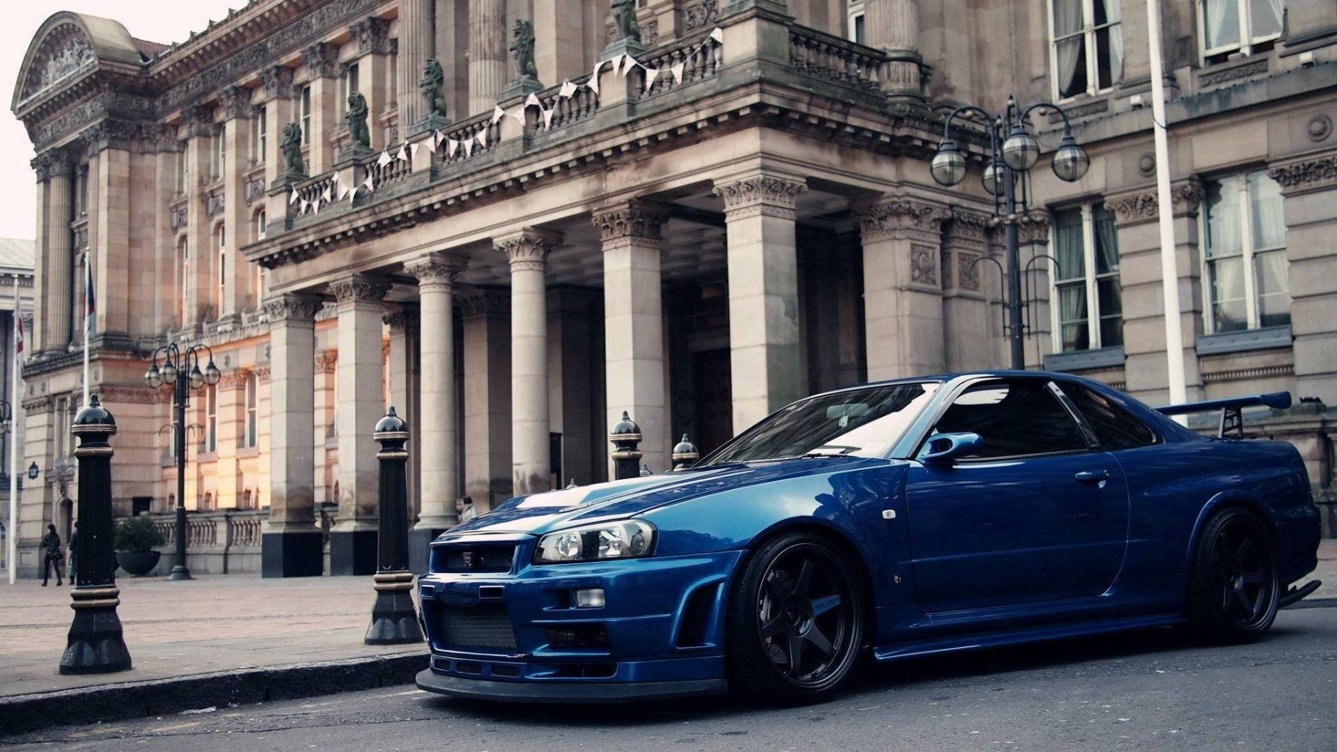 天際線 桌布 壁紙 — 一輛光亮的藍色日產Skyline R34跑車停在一棟帶有柱子的宏偉古典石建築前。