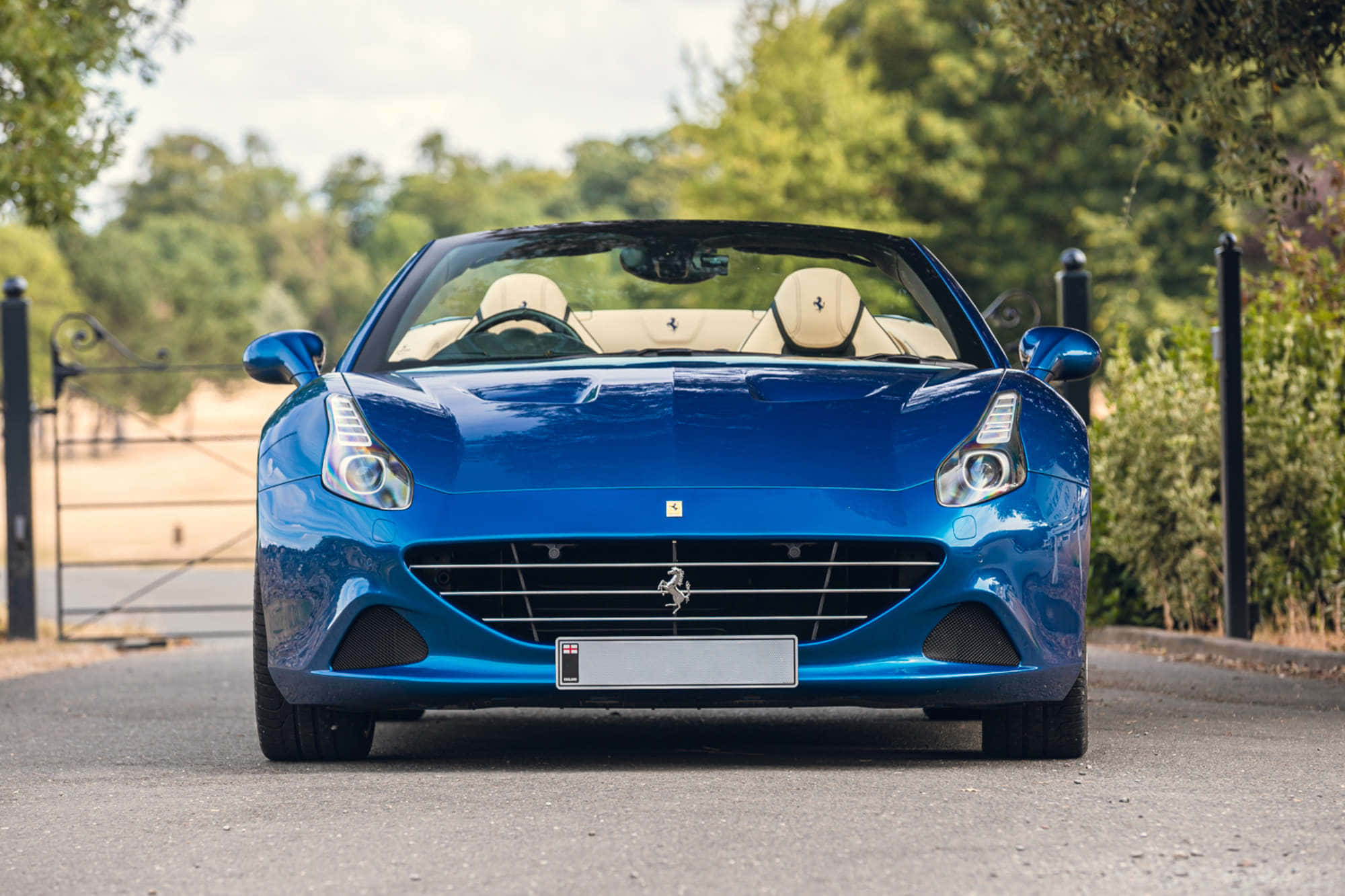 Ferrari California T 桌布 壁紙 — 一輛光亮的藍色法拉利敞篷車停在鋪砌的道路上，背景有茂密的綠樹和一個鍛鐵大門。