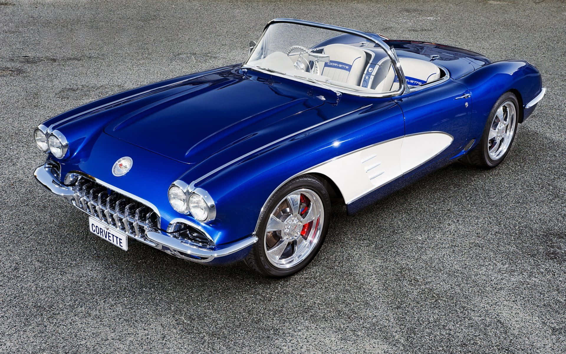 classic 桌布 壁紙 — 一輛光亮的藍色經典Corvette敞篷車，帶有白色點綴，停在灰色瀝青上，在明亮的日光下。