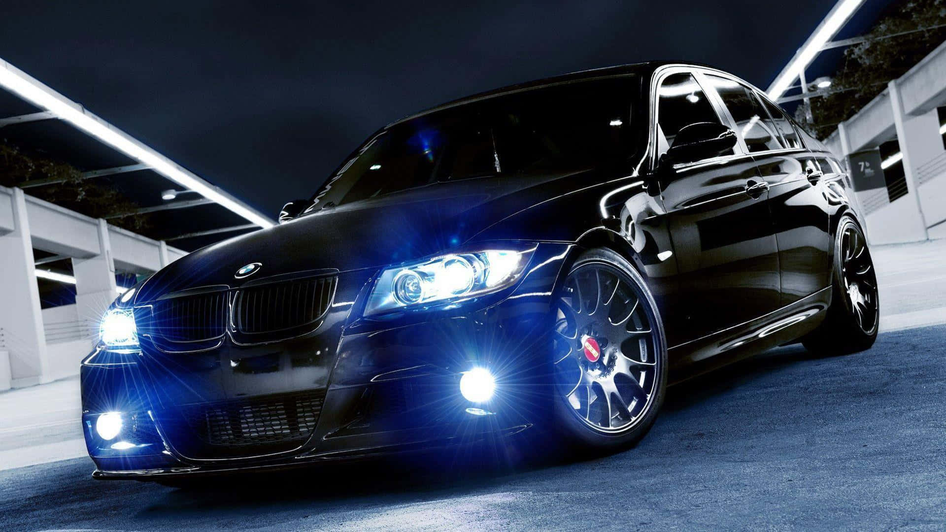 Cool Bmw 桌布 壁紙 — 一輛光亮的黑色寶馬轎車在夜間停放在現代停車場的明亮天花燈下，車頭燈閃爍發光。
