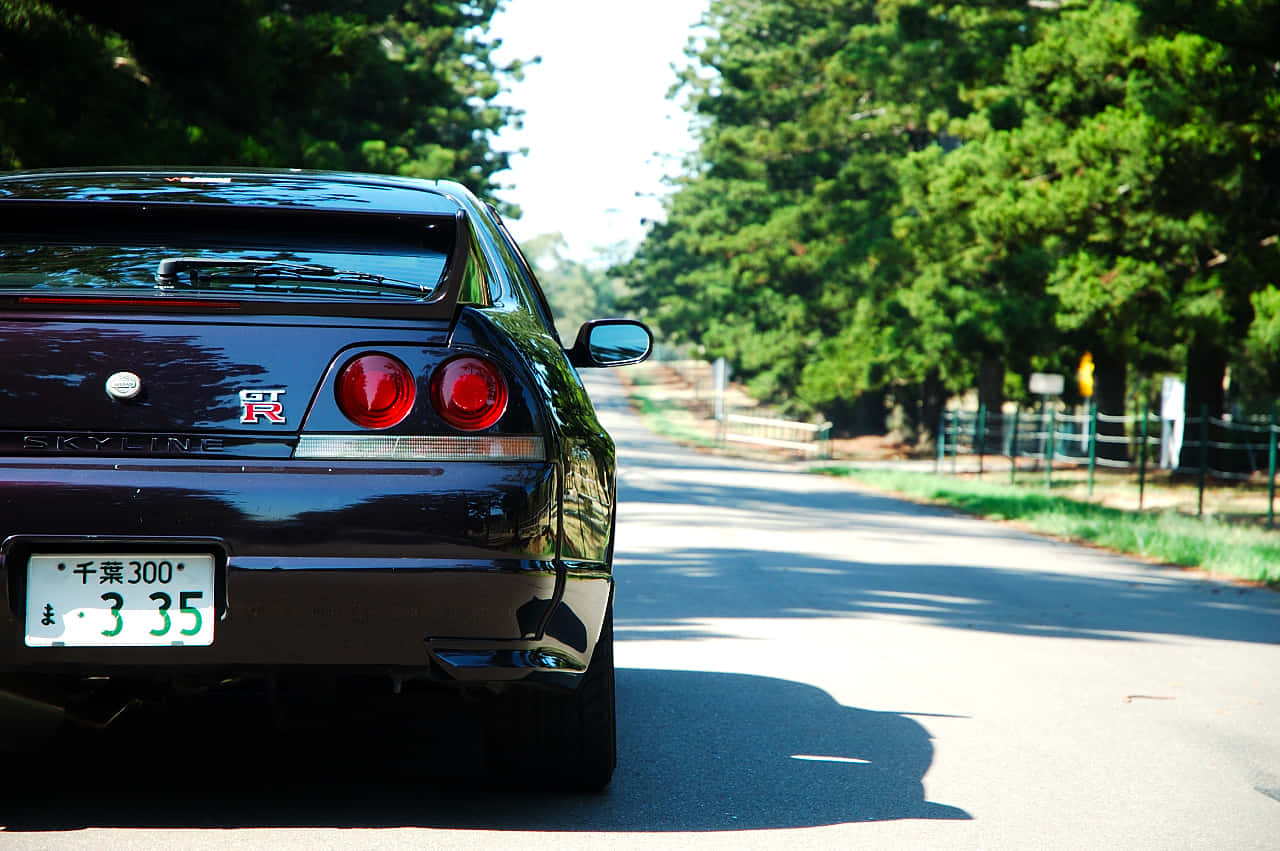 R33 GT-R 桌布 壁紙 — 一輛光亮的黑色日產Skyline R34跑車停泊在陽光普照、綠樹成蔭的道路上，投下清晰的影子。