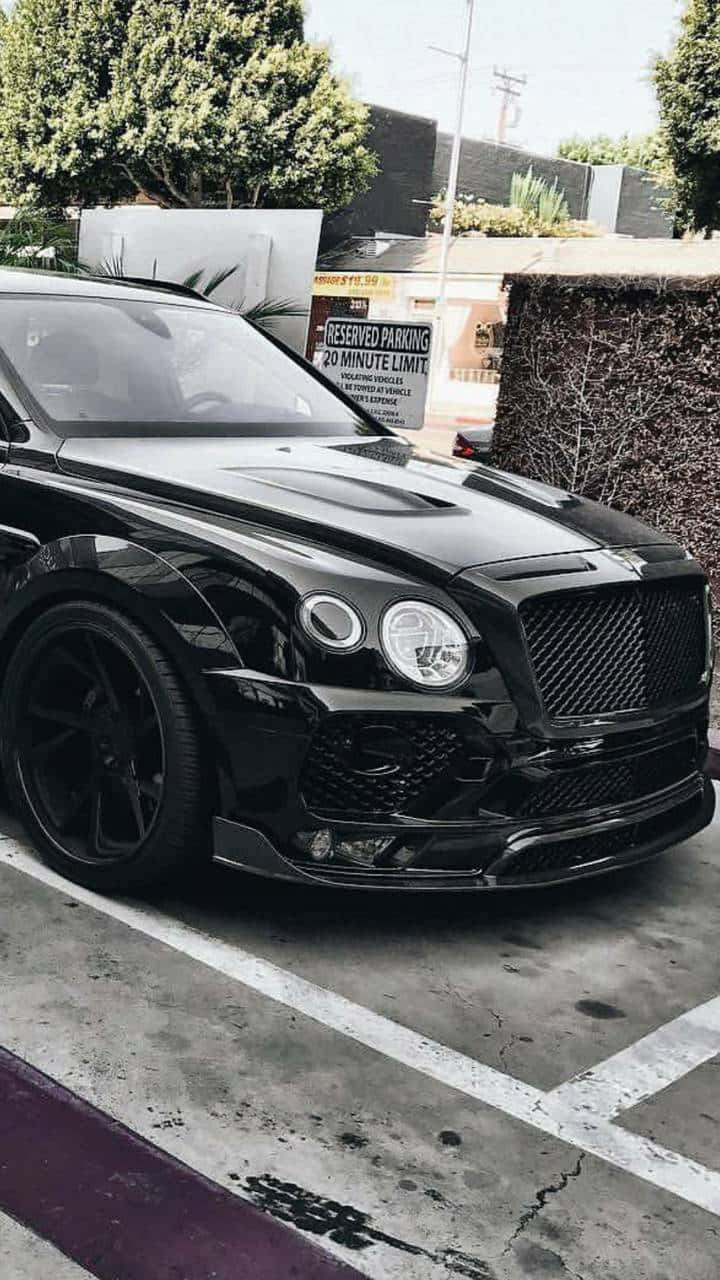 汽車 iPhone 壁紙 桌布 壁紙 — 一輛光亮的黑色賓利SUV，配備定制輪圈，停放在陽光明媚的城市地段，旁邊有樹籬和預留停車標誌。