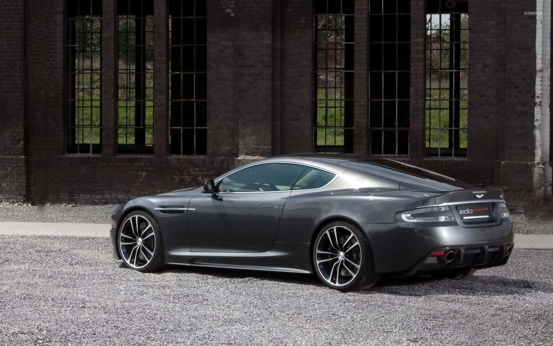 Aston Martin Db9 桌布 壁紙 — 一輛光滑的啞光灰色 Aston Martin 跑車停泊在碎石路上，前面是一棟帶有高窗的風化磚砌建築。