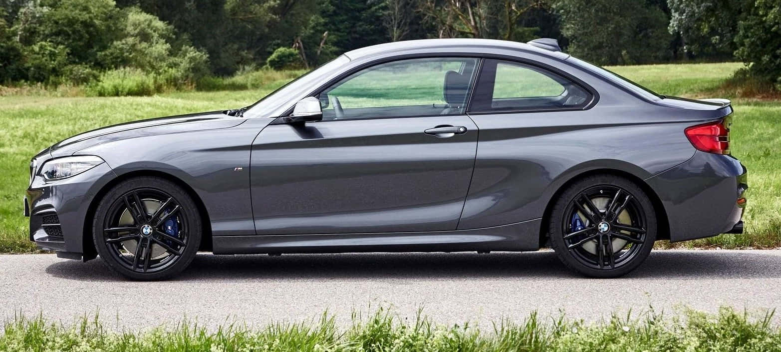 Bmw 2 Series 桌布 壁紙 — 一輛光滑的啞光灰色寶馬雙門轎跑車停泊在碎石路上，旁邊是充滿活力的綠草和樹木。