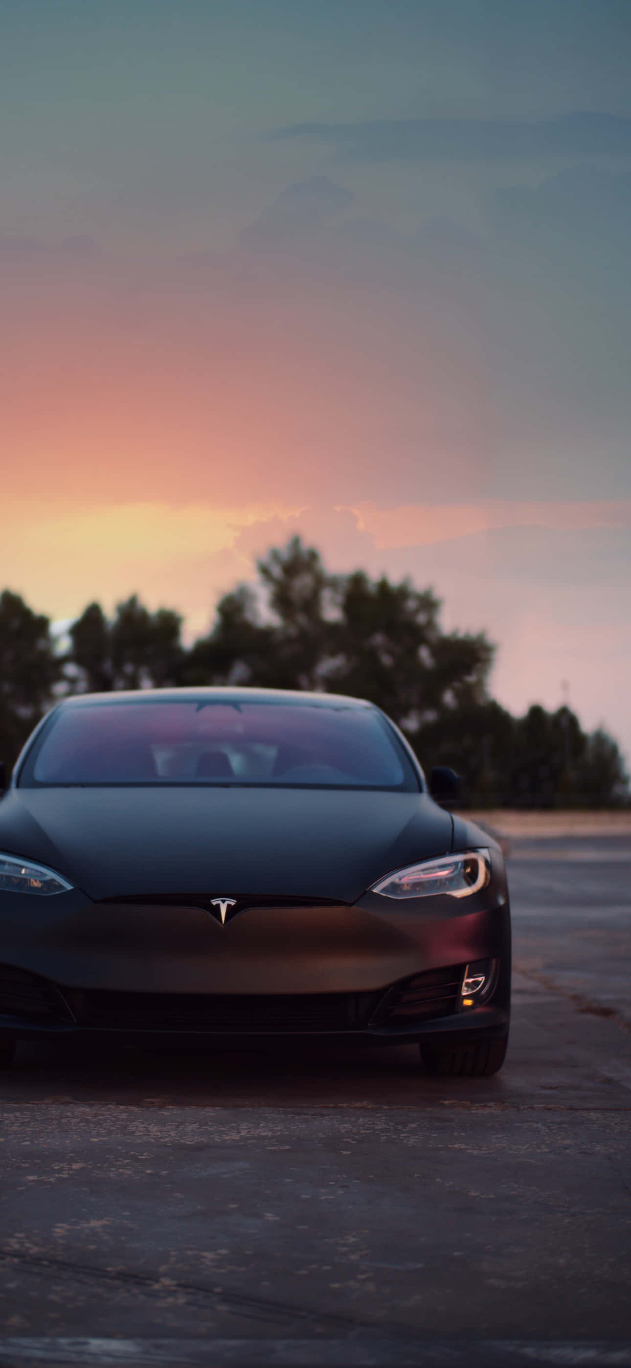 Tesla iPhone 桌布 壁紙 — 一輛光滑的啞光黑色Tesla停在瀝青路上，背景是充滿活力的日落天空和剪影樹木。