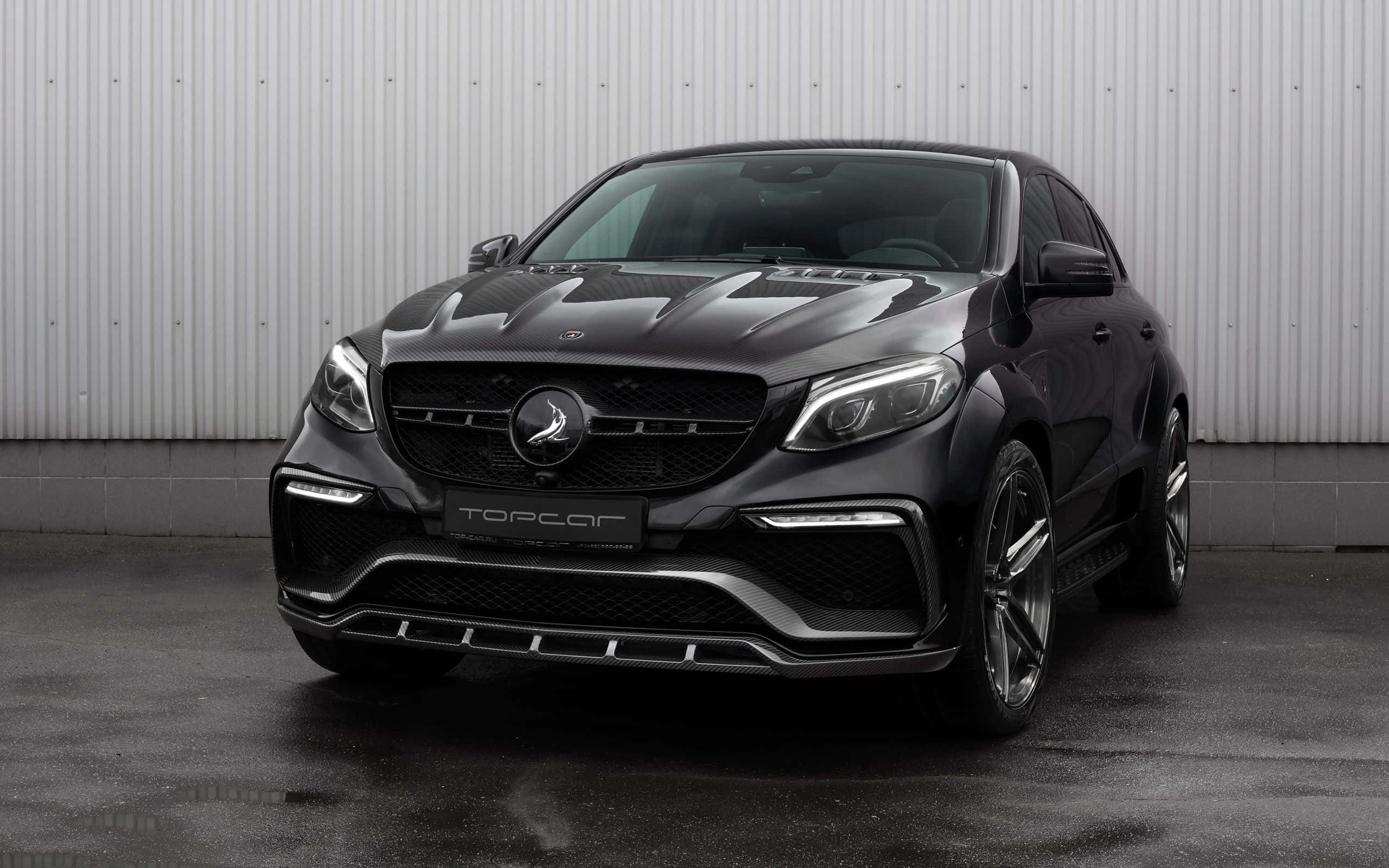 桌布 壁紙 — 一輛光滑的啞光黑色改裝梅賽德斯-AMG GLE Coupe，帶有碳纖維飾件和定製「TOPCAR」徽章，停靠在波紋金屬牆旁。