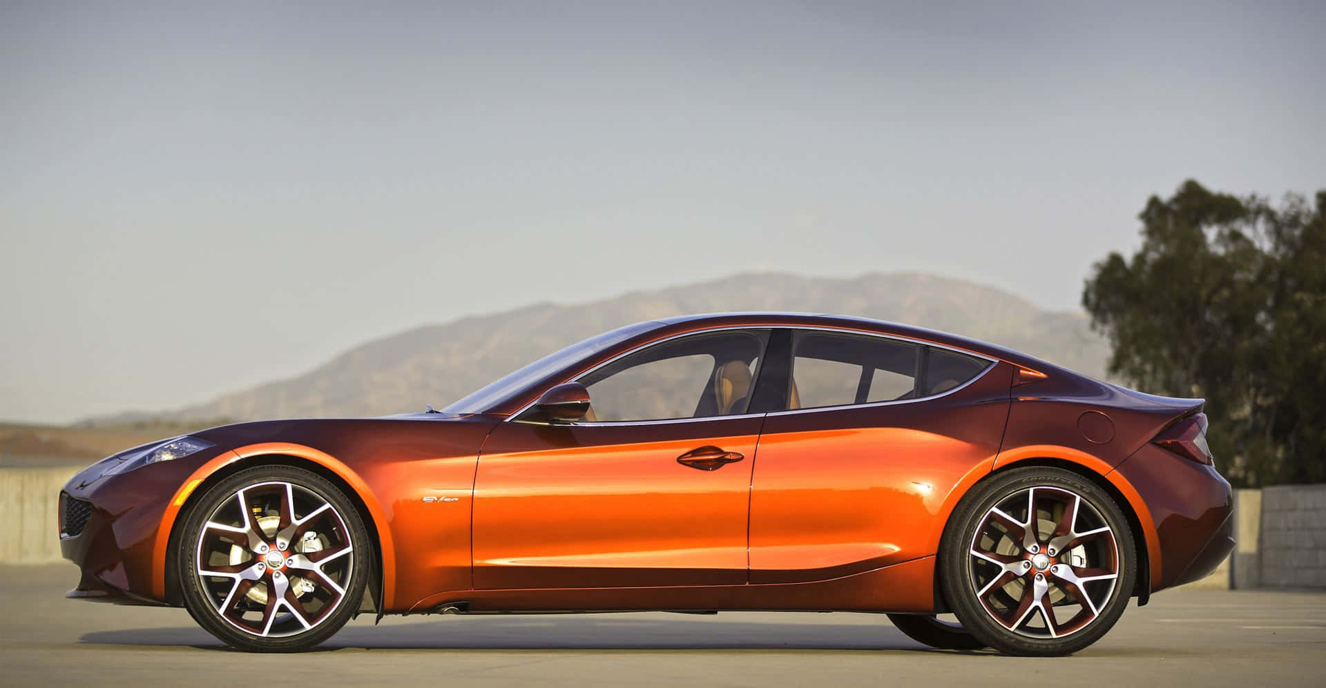 Fisker 桌布 壁紙 — 一輛光滑的橙色跑車，帶有光澤飾面，停泊在鋪砌的路面上，背景是山脈和樹木，天空晴朗。