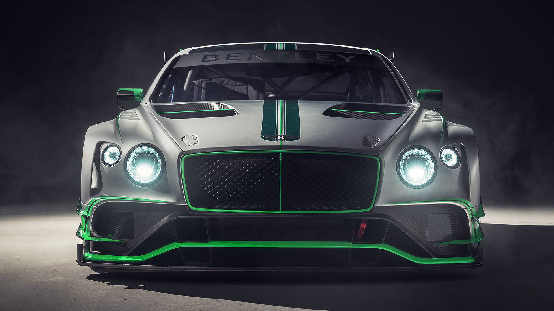 bentley HD 桌布 壁紙 — 一輛光滑的銀色賓利賽車，帶有發光的綠色飾邊和車頭燈，在黑暗的攝影棚環境中凝視前方。