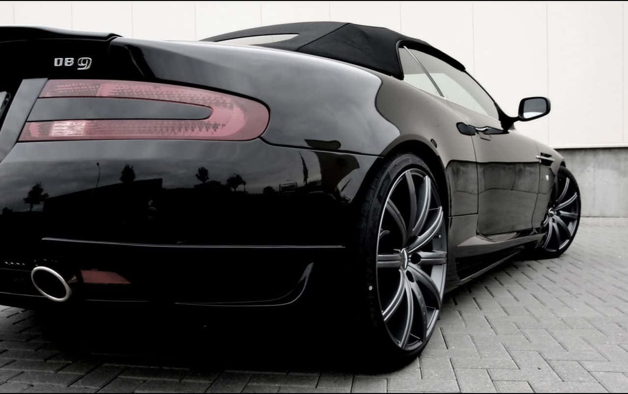Aston Martin Db9 桌布 壁紙 — 一輛光滑的黑色Aston Martin DB9敞篷跑車停放在鋪砌路面上，其光澤表面反射著周圍環境。