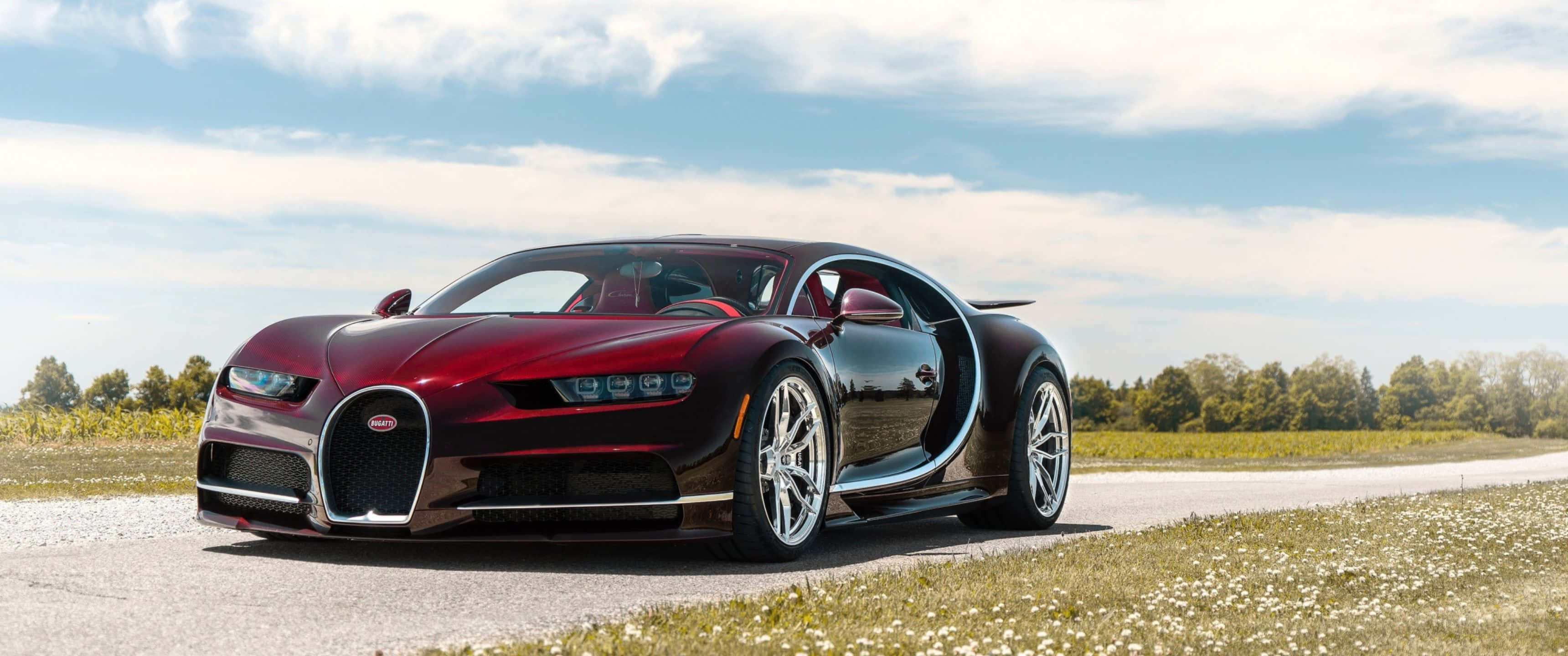 3440x1440p Bugatti 桌布 壁紙 — 一輛光澤的栗色Bugatti Chiron打開剪刀門，停在碎石路上，旁邊是野花田，天空部分多雲。