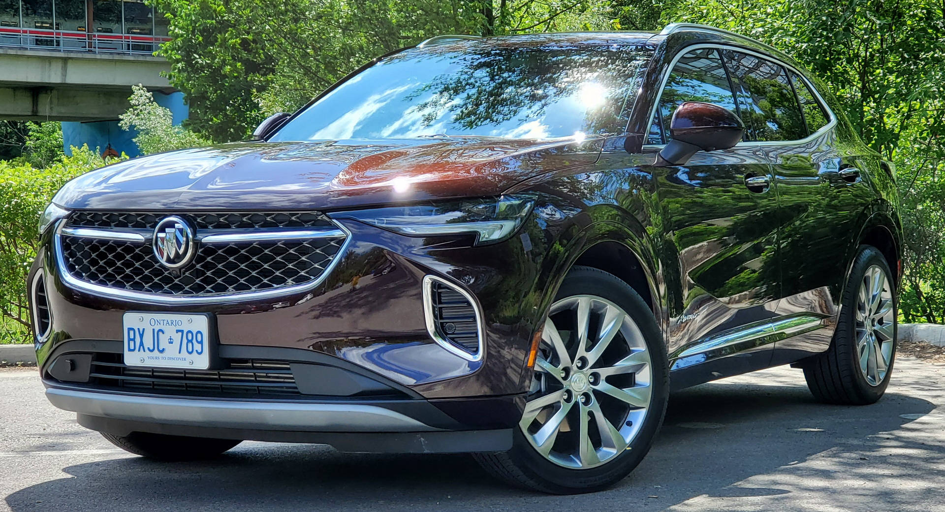 Buick 桌布 壁紙 — 一輛光澤的栗色Buick SUV停放在戶外，在明亮的陽光下，背景是茂密的綠樹和一座橋樑。
