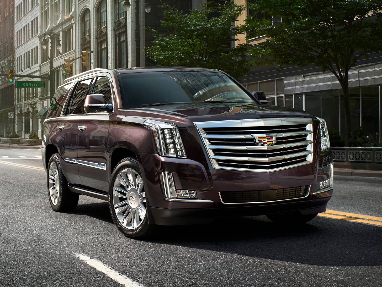 凱迪拉克 iPhone 桌布 壁紙 — 一輛光澤的栗色Cadillac Escalade SUV停放在陽光普照的城市街道上，背景是歷史建築和綠樹。