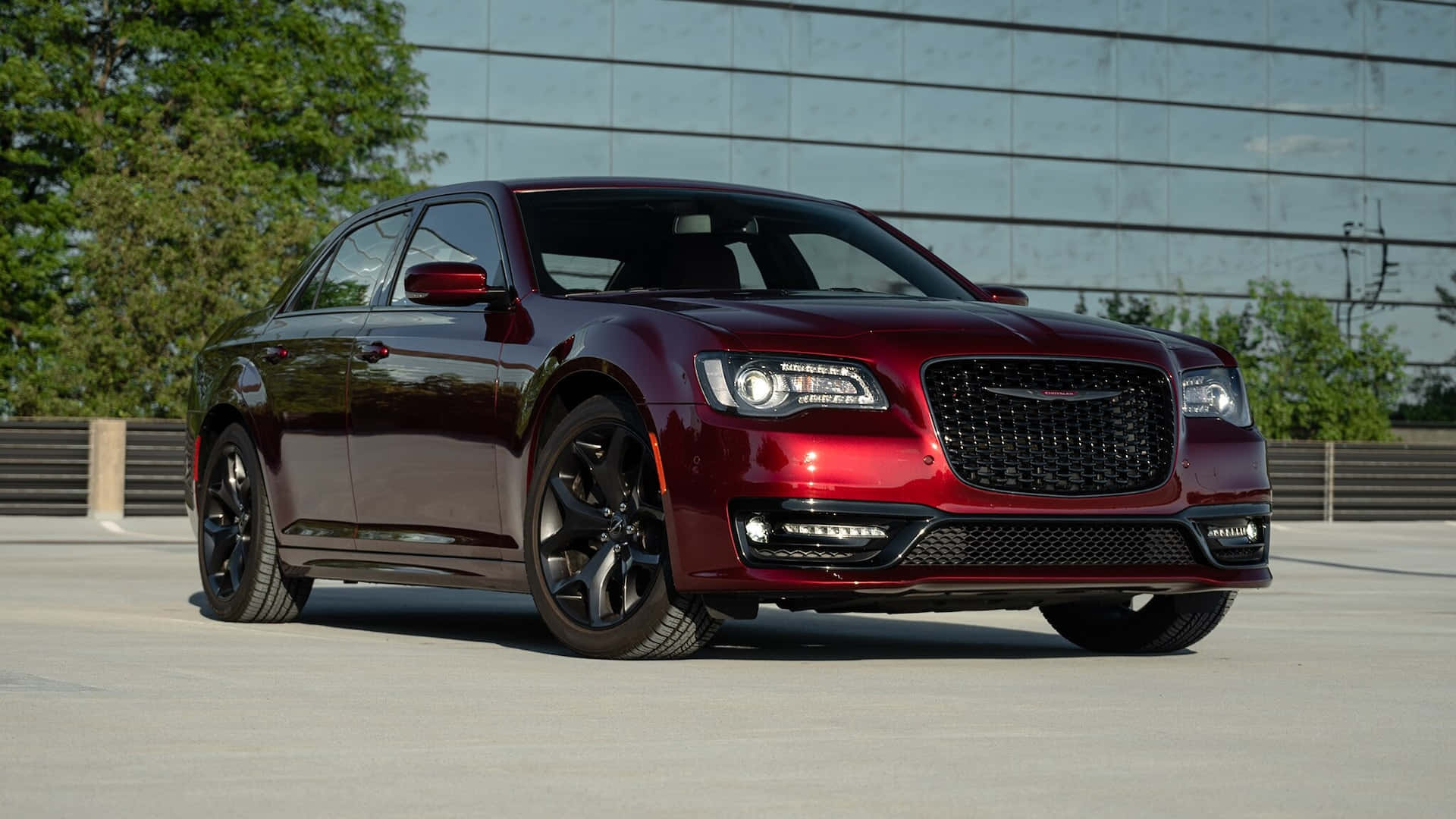 克萊斯勒 桌布 壁紙 — 一輛光澤的栗色Chrysler 300轎車，搭配黑色輪圈，停在混凝土停車場上，背景是玻璃建築和綠樹。