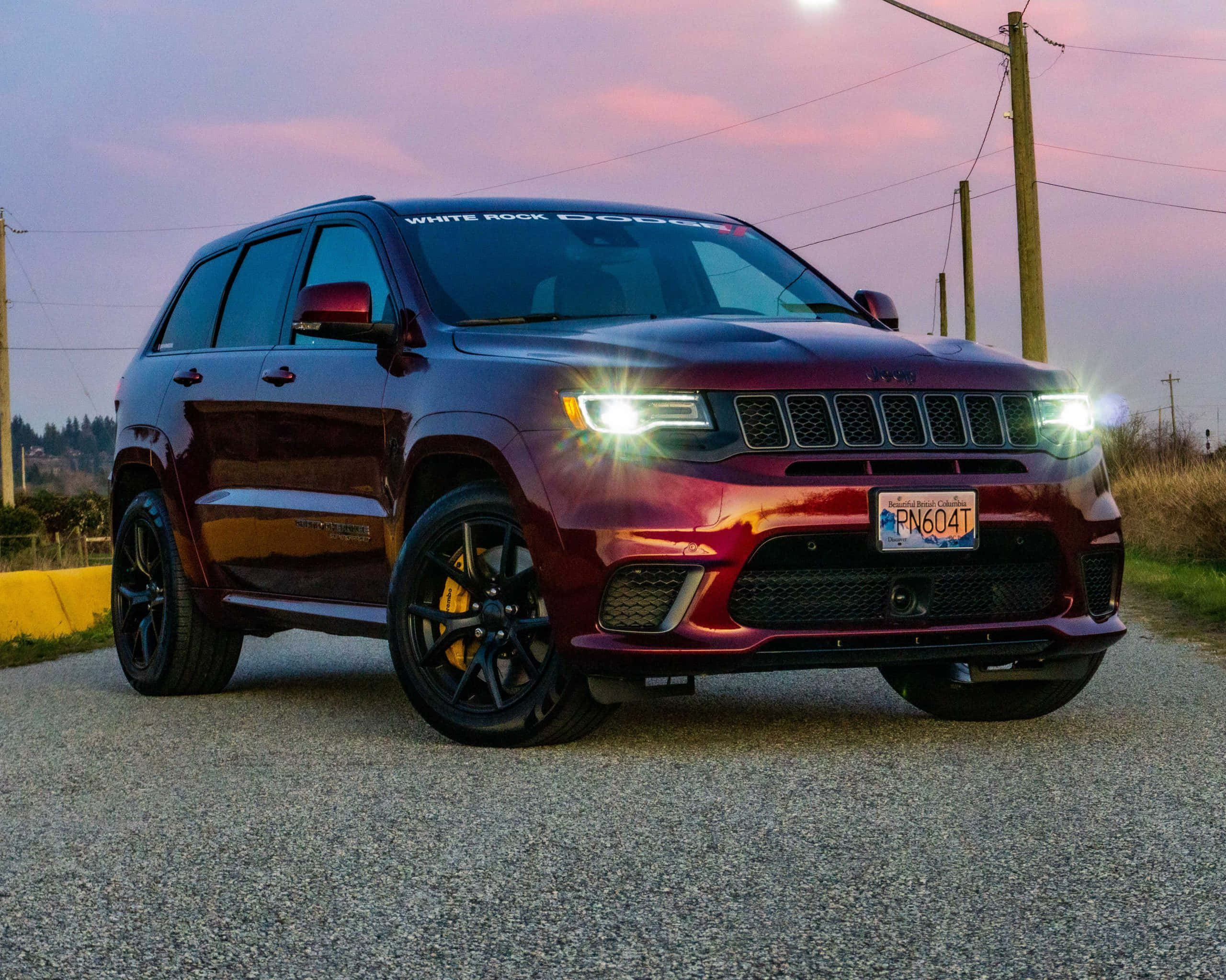 Jeep Trackhawk 桌布 壁紙 — 一輛光澤的栗色Jeep Grand Cherokee Trackhawk，配備黑色輪圈和黃色制動卡鉗，在黃昏時停泊在礫石上，車頭燈亮著。