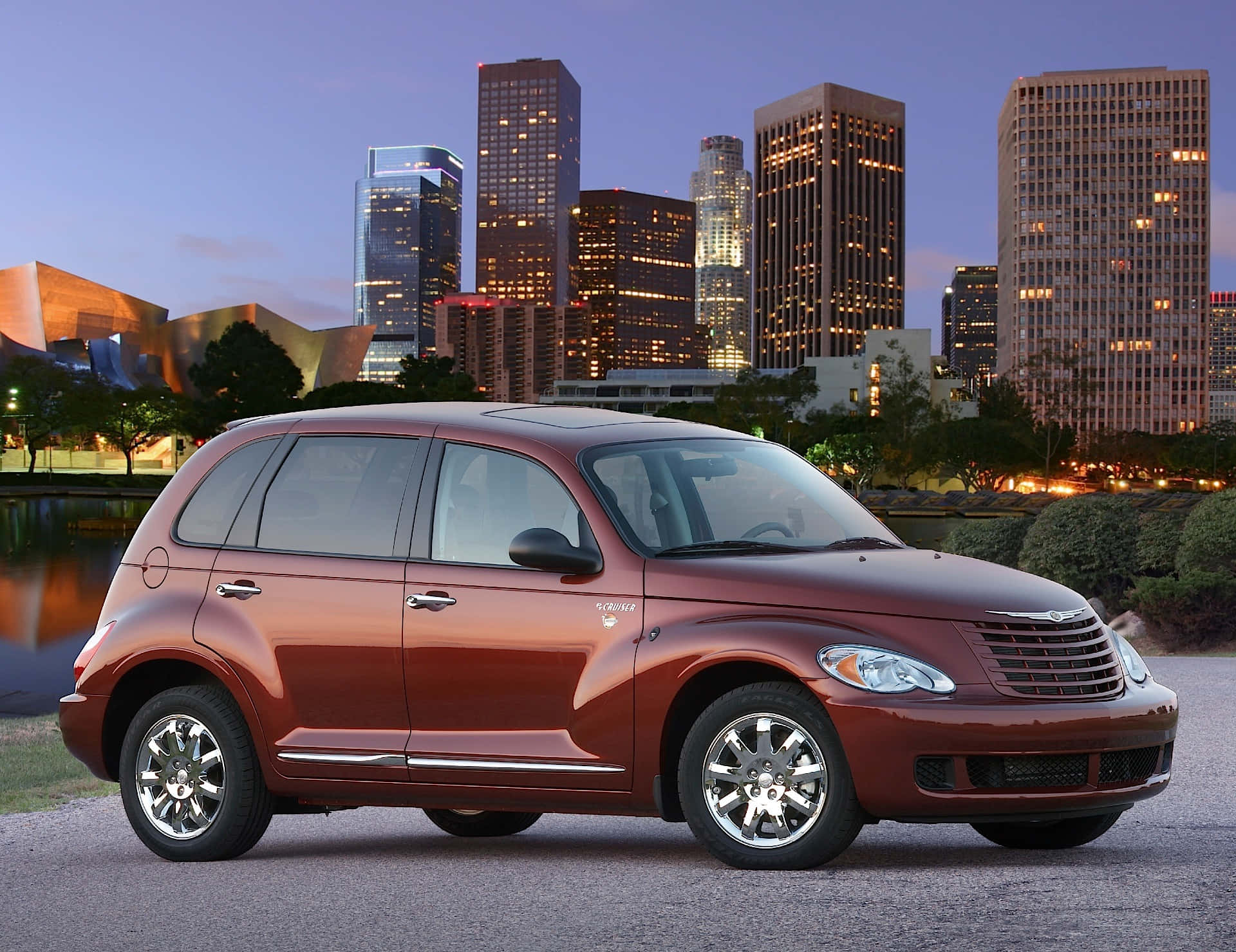 Chrysler Pt Cruiser 桌布 壁紙 — 一輛光澤的栗色PT Cruiser停在人行道上，背後是暮色中的城市天際線和亮燈的摩天大樓。