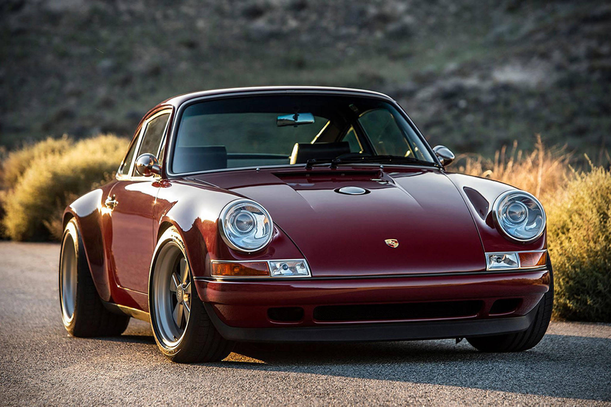 Singer 桌布 壁紙 — 一輛光澤的栗色保時捷911經典跑車停泊在碎石路上，旁邊有乾燥灌木，沐浴在溫暖的金色陽光下。