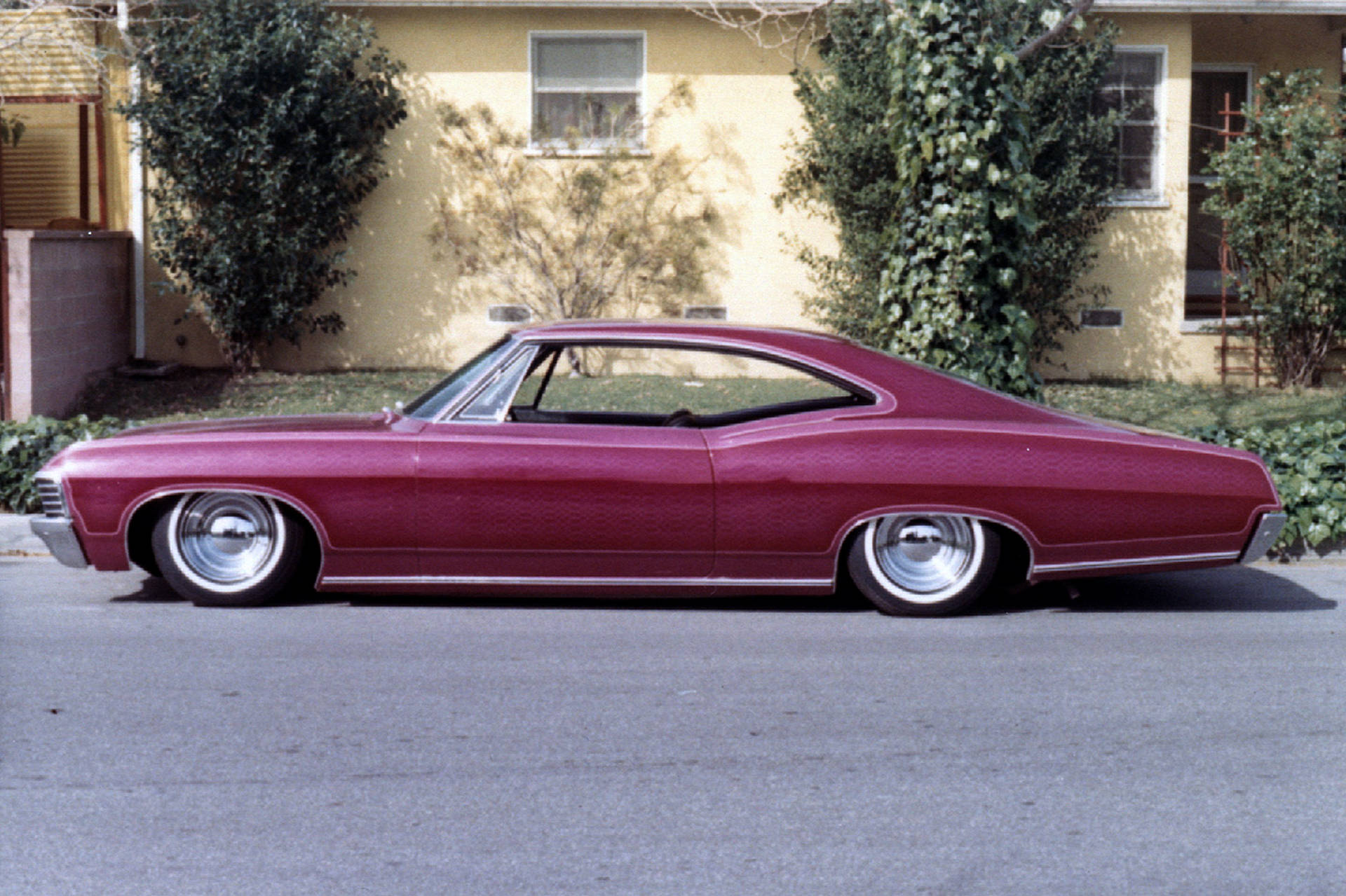 chevrolet impala 1967 桌布 壁紙 — 一輛光澤的栗色經典肌肉車，配有鍍鉻輪圈，停在黃色房子前的街道上，房子周圍有綠色灌木。