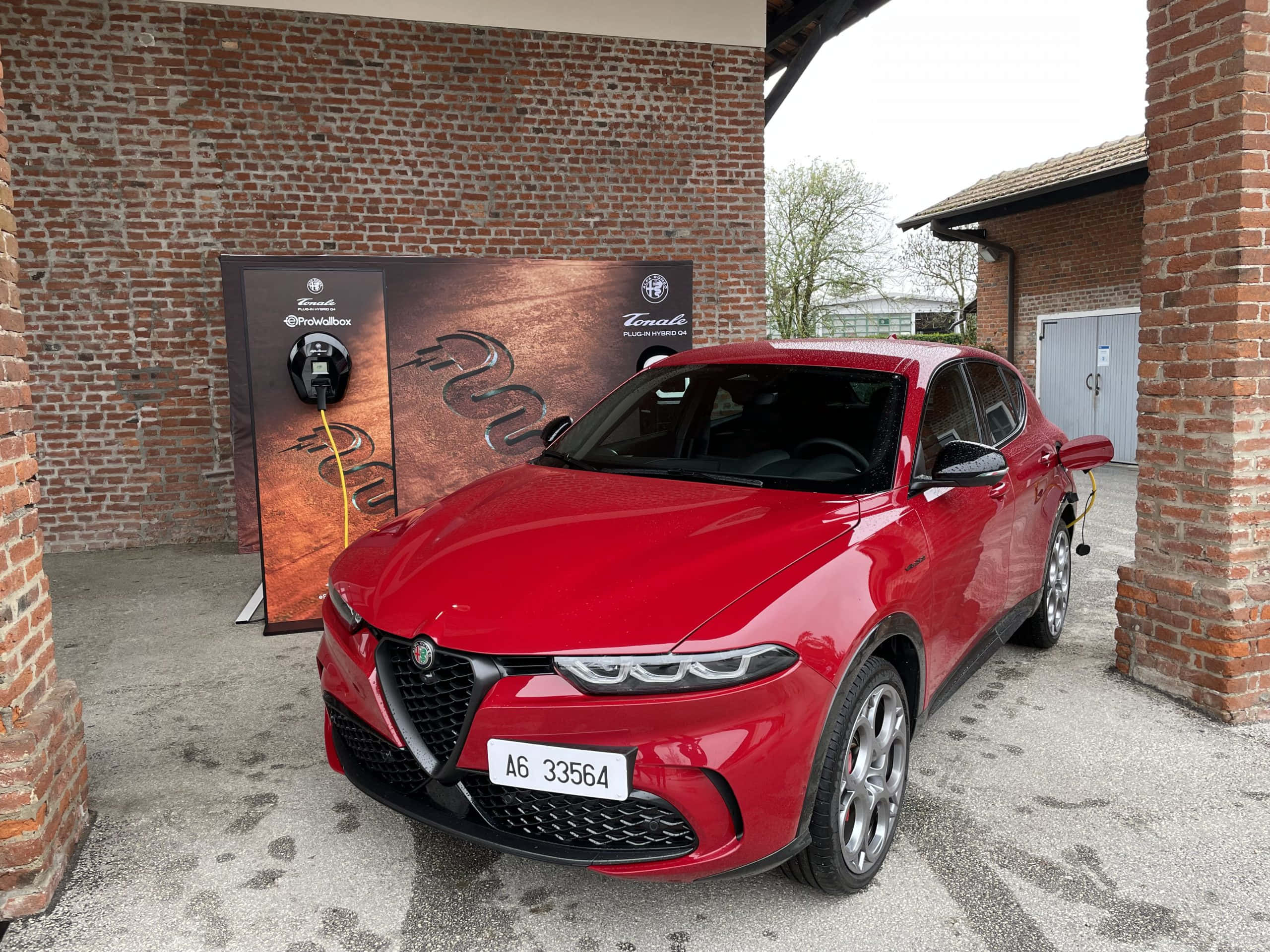 愛快羅密歐 桌布 壁紙 — 一輛光澤的紅色 Alfa Romeo Tonale 插電式混合動力 SUV 在有蓋天空下，在磚砌建築旁的品牌充電站充電。