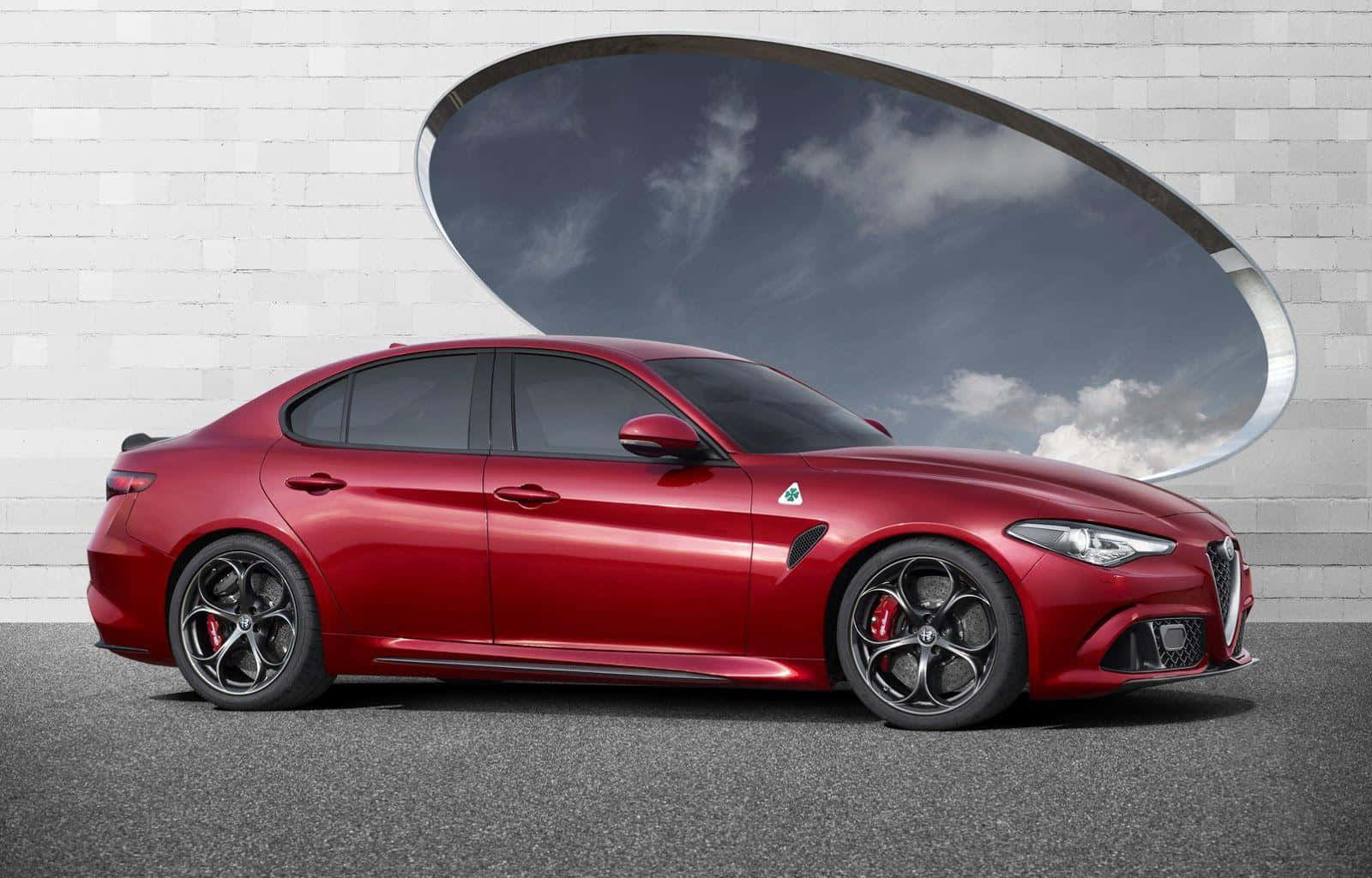愛快羅密歐 桌布 壁紙 — 一輛光澤的紅色 Alfa Romeo 轎車停放在白色磚牆前，牆上有一個大橢圓形窗戶，框著多雲的天空。
