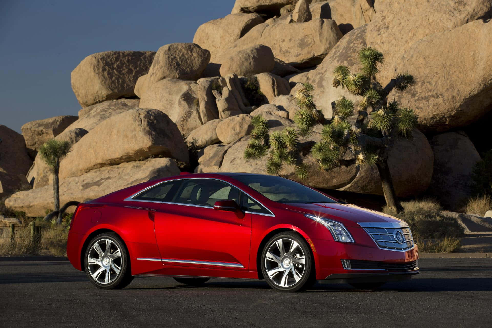 Cadillac Elr 桌布 壁紙 — 一輛光澤的紅色 Cadillac ELR 雙門轎跑車在強烈的陽光下閃閃發光，旁邊是巨大的沙漠巨石和約書亞樹。