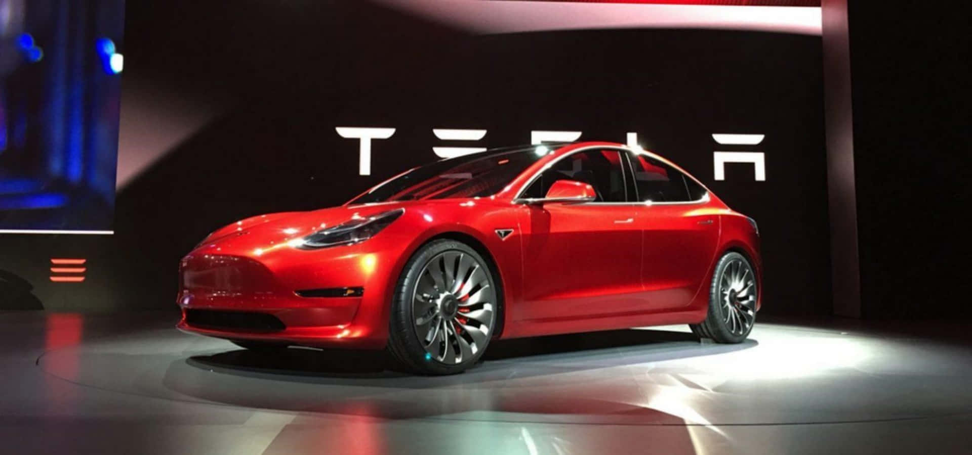 Tesla Model 3 桌布 壁紙 — 一輛光澤的紅色 Tesla Model 3 轎車在明亮的聚光燈下於圓形舞台上展示，背後 Tesla 標誌閃閃發光。