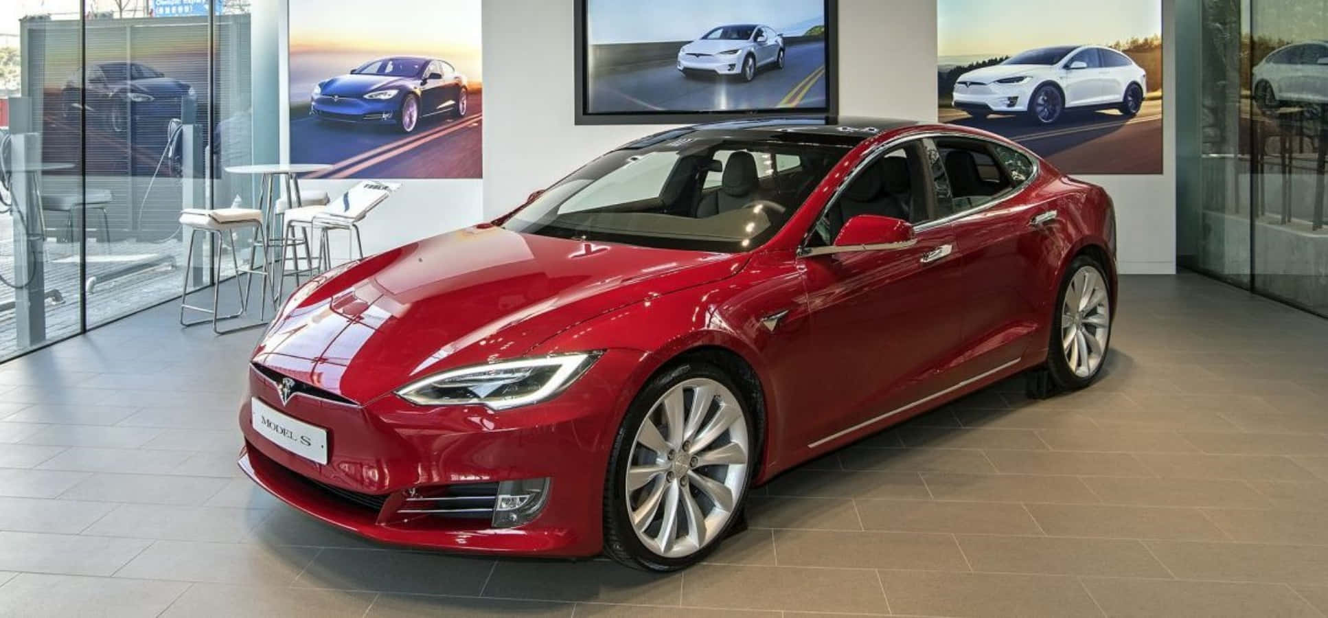 特斯拉 桌布 壁紙 — 一輛光澤的紅色 Tesla Model S 轎車在現代汽車經銷商展廳中展示，展廳有大型玻璃窗和牆上的促銷海報。