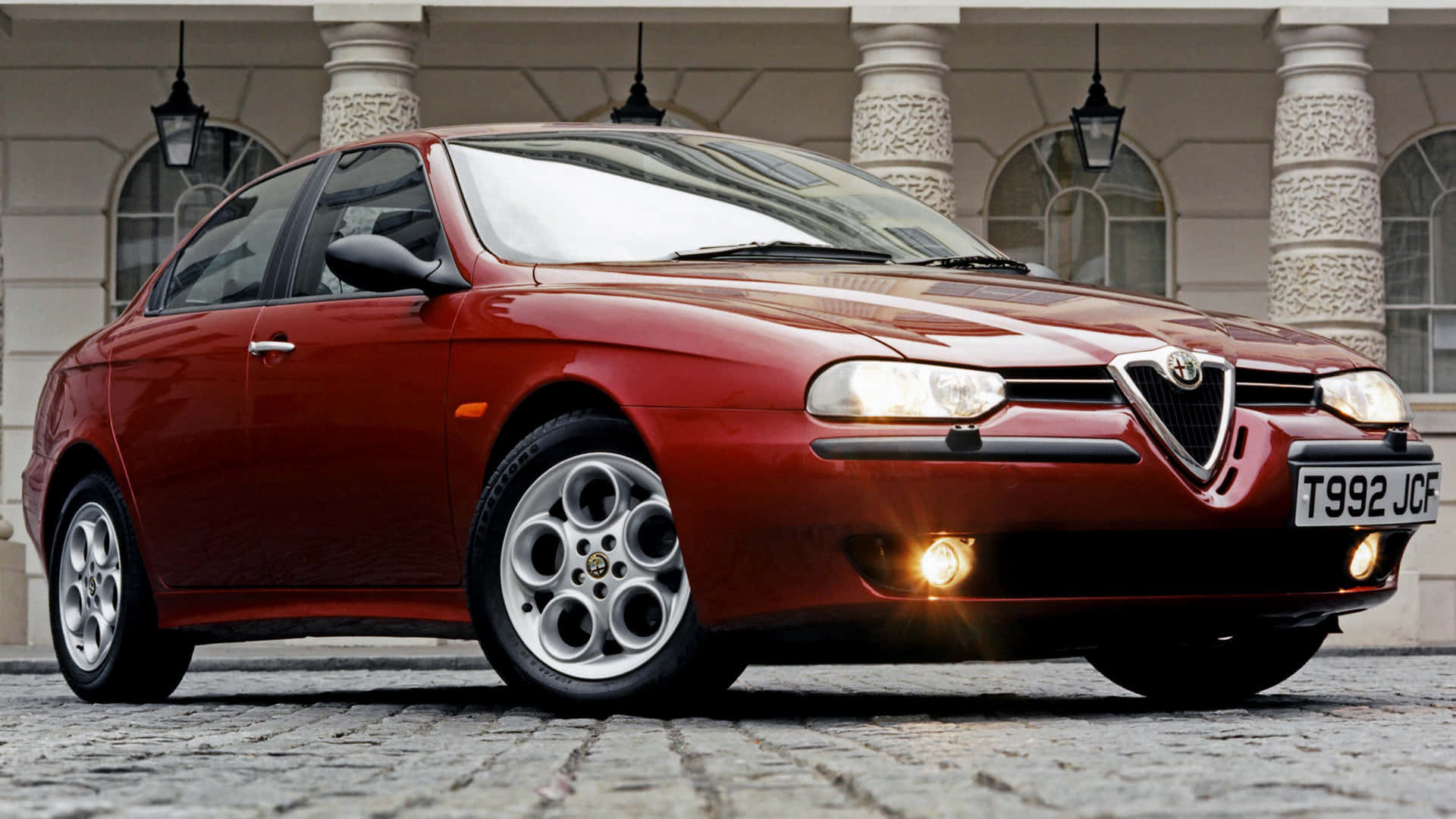 Alfa Romeo 156 桌布 壁紙 — 一輛光澤的紅色Alfa Romeo轎車停在鵝卵石街道上，前方是帶有華麗柱子的古典建築。
