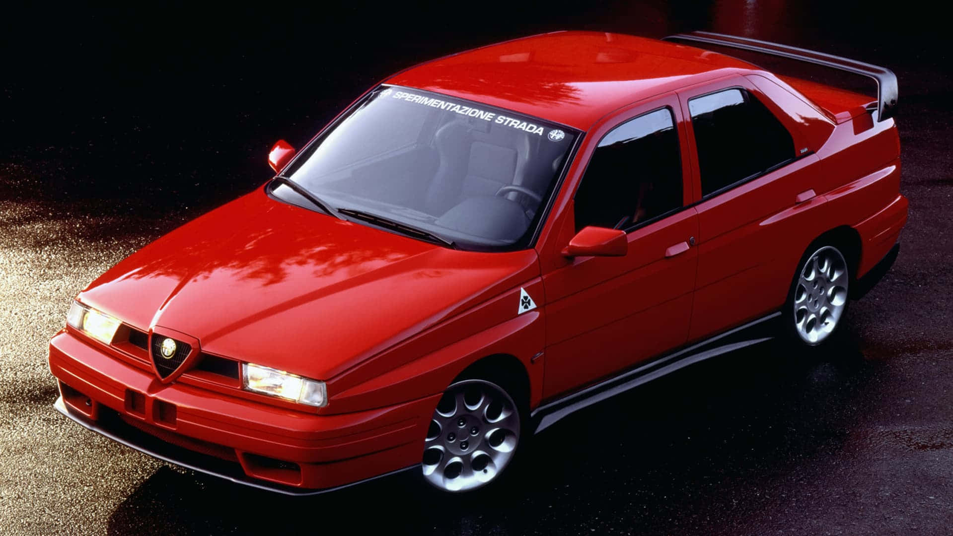 愛快羅密歐 桌布 壁紙 — 一輛光澤的紅色Alfa Romeo 155 V6 Sport轎車在潮濕的瀝青路面上於攝影棚燈光下閃耀。