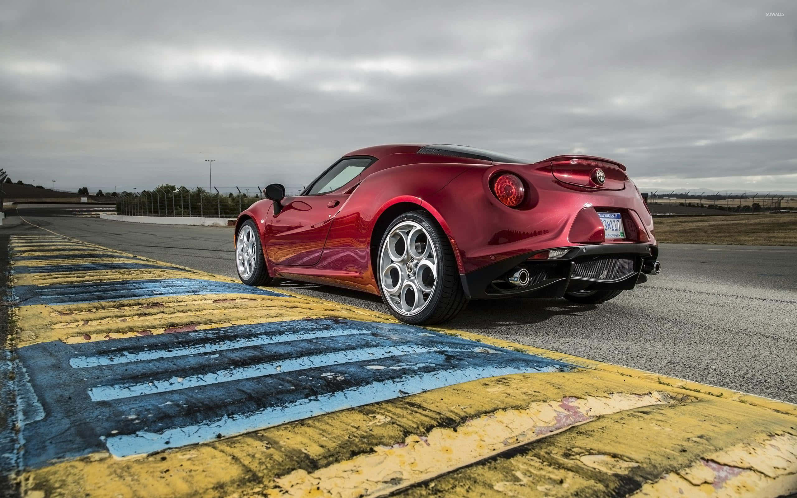 Alfa Romeo 4c 桌布 壁紙 — 一輛光澤的紅色Alfa Romeo 4C跑車停泊在風化的黃藍色賽道邊緣，天空多雲。