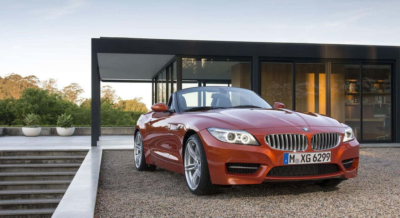 BMW 動圖 桌布 壁紙 — 一輛光澤的紅色BMW Z4敞篷跑車停泊在碎石路上，前方是一座現代玻璃屋，室內燈光溫暖。