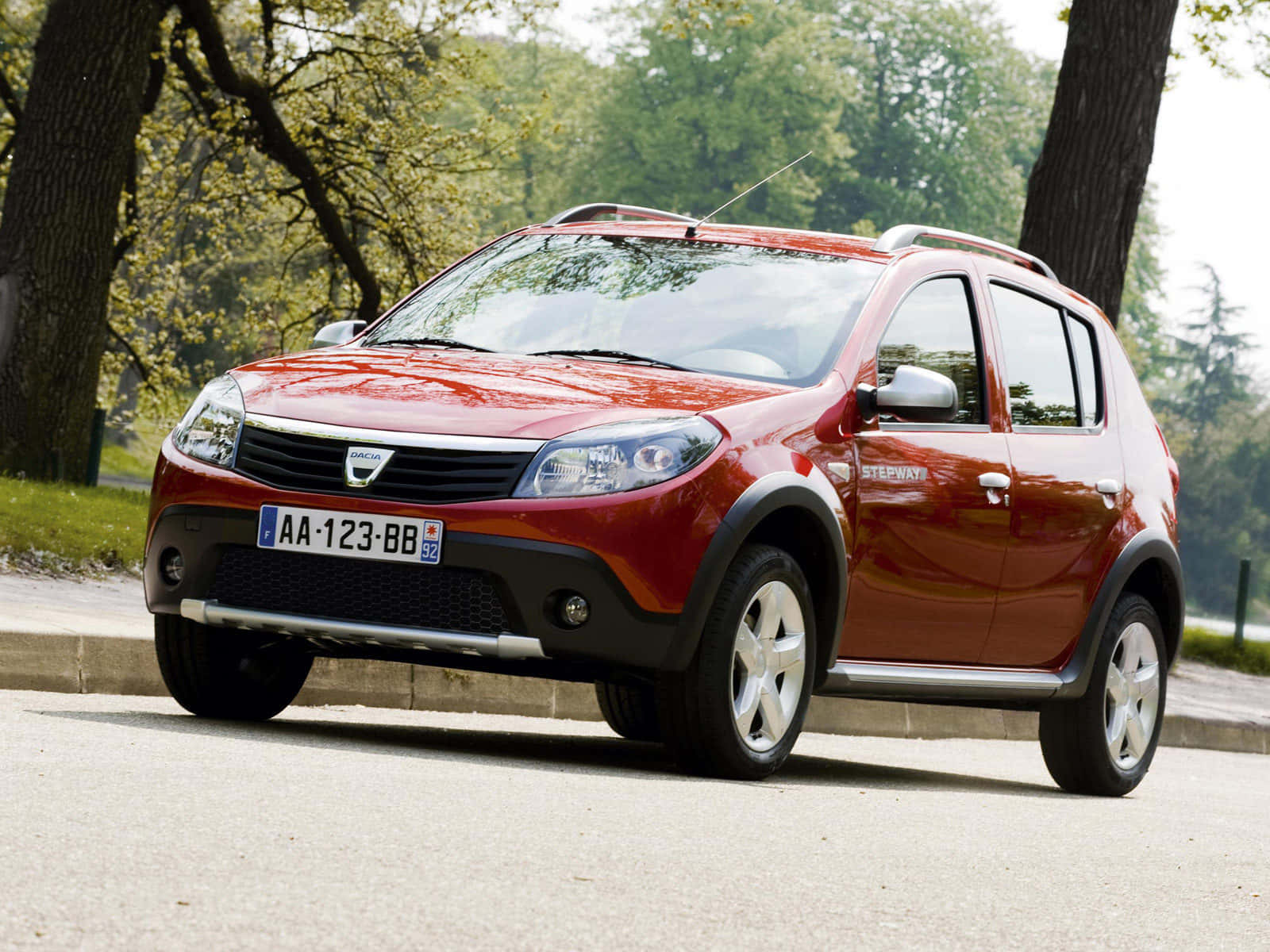 Dacia 桌布 壁紙 — 一輛光澤的紅色Dacia Stepway SUV停放在綠樹成蔭的街道上，柔和日光下綠葉繁茂。