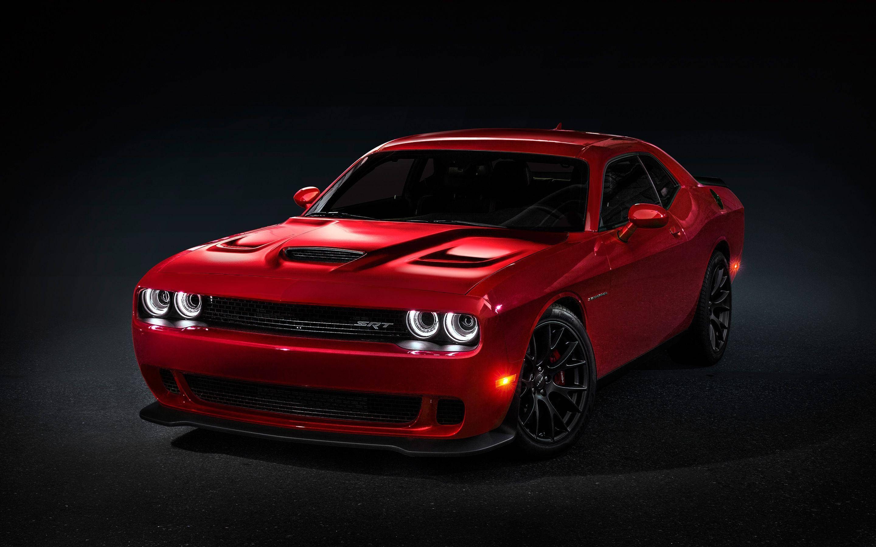 Challenger 桌布 壁紙 — 一輛光澤的紅色Dodge Challenger SRT，帶有發光的頭燈和黑色輪圈，在黑暗的攝影棚背景下戲劇性地照亮。