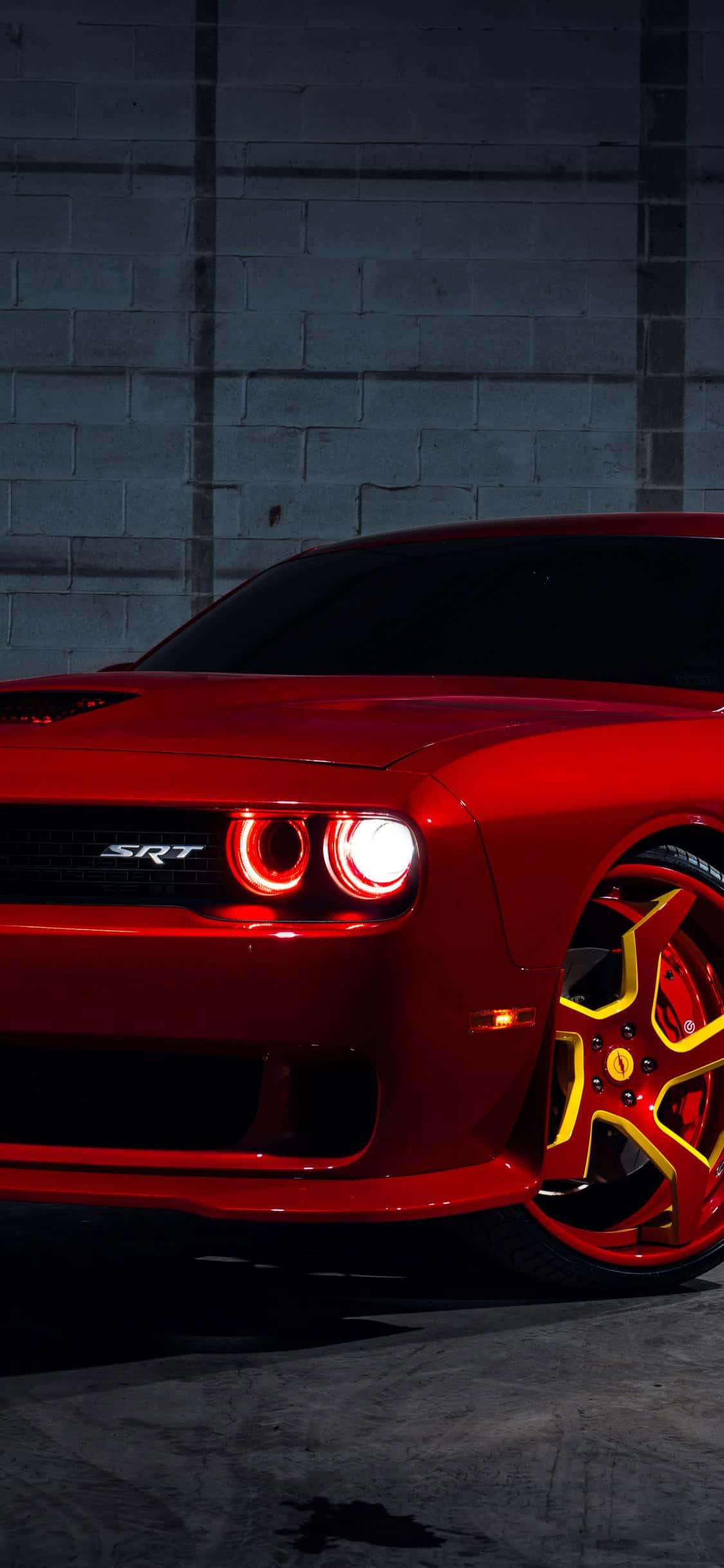 桌布 壁紙 — 一輛光澤的紅色Dodge Challenger SRT，帶有發光的頭燈和黃色點綴的輪圈，停在深色混凝土牆前。
