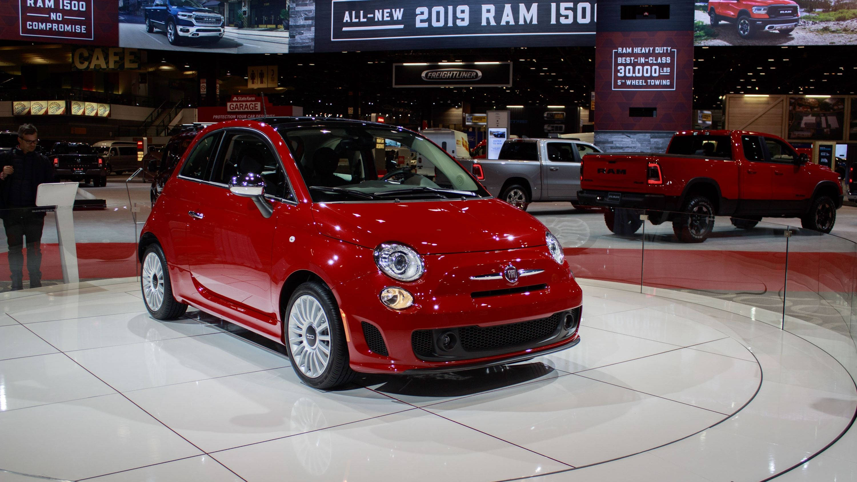 Fiat 桌布 壁紙 — 一輛光澤的紅色Fiat 500在圓形展廳地板上，背景有其他卡車和促銷橫幅。