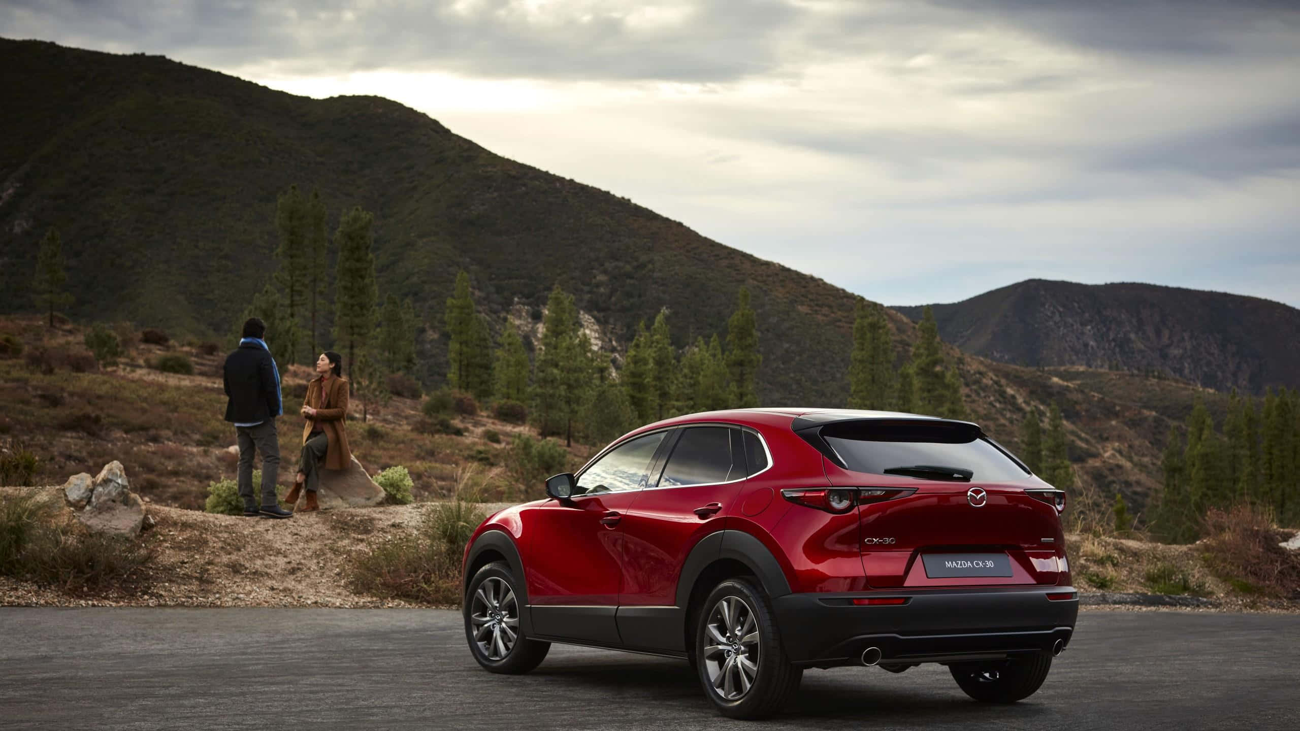 Mazda Cx-30 桌布 壁紙 — 一輛光澤的紅色Mazda CX-30 SUV停放在鋪砌道路上，附近站著一對情侶，背景是起伏的山丘和稀疏的樹木，天空多雲。