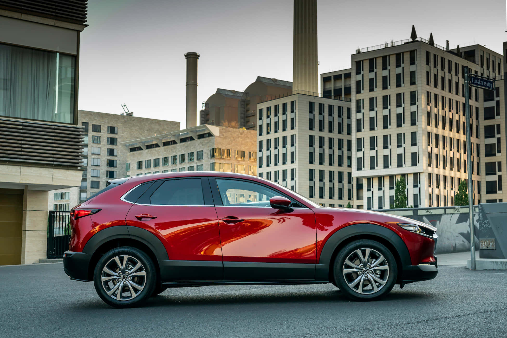 Mazda Cx-30 桌布 壁紙 — 一輛光澤的紅色Mazda CX-30 SUV在黃昏時停泊在現代建築和高煙囪背景的城市街道上。