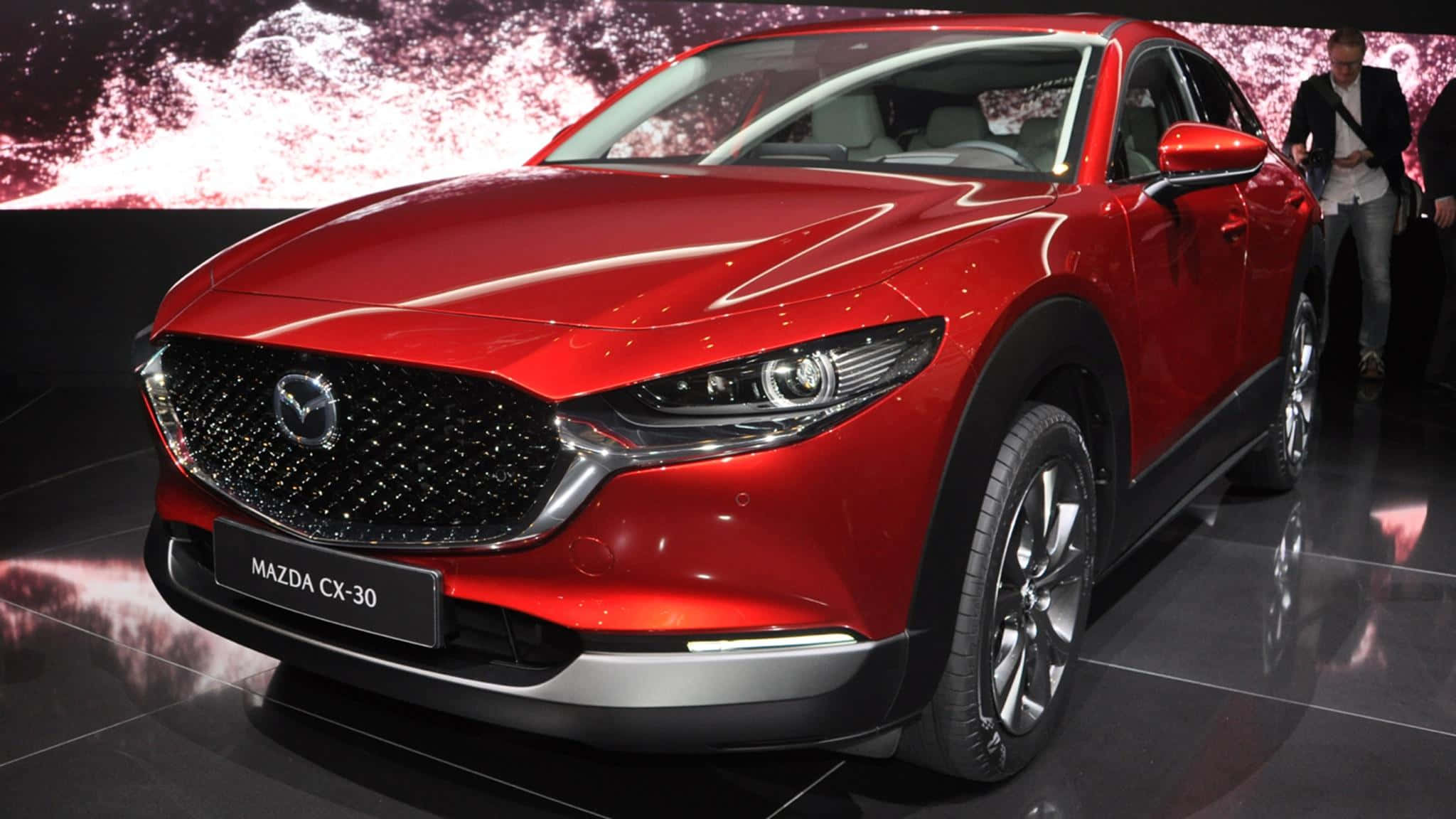 Mazda Cx-30 桌布 壁紙 — 一輛光澤的紅色Mazda CX-30 SUV在車展上明亮的燈光下閃耀，背景動感抽象。