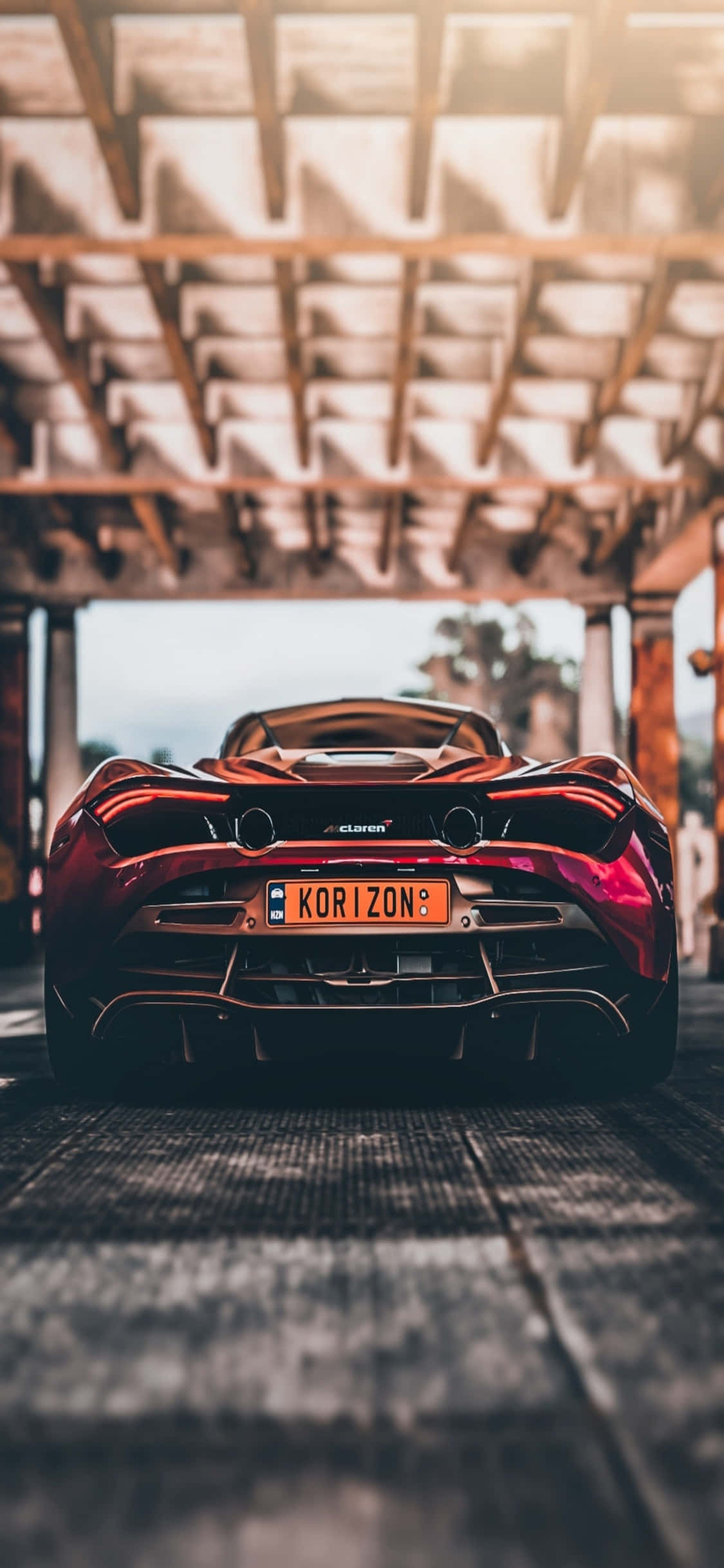 iPhone Xs Mclaren 720s 桌布 壁紙 — 一輛光澤的紅色McLaren跑車停放在木結構下，陽光透過上方的橫樑照射進來。
