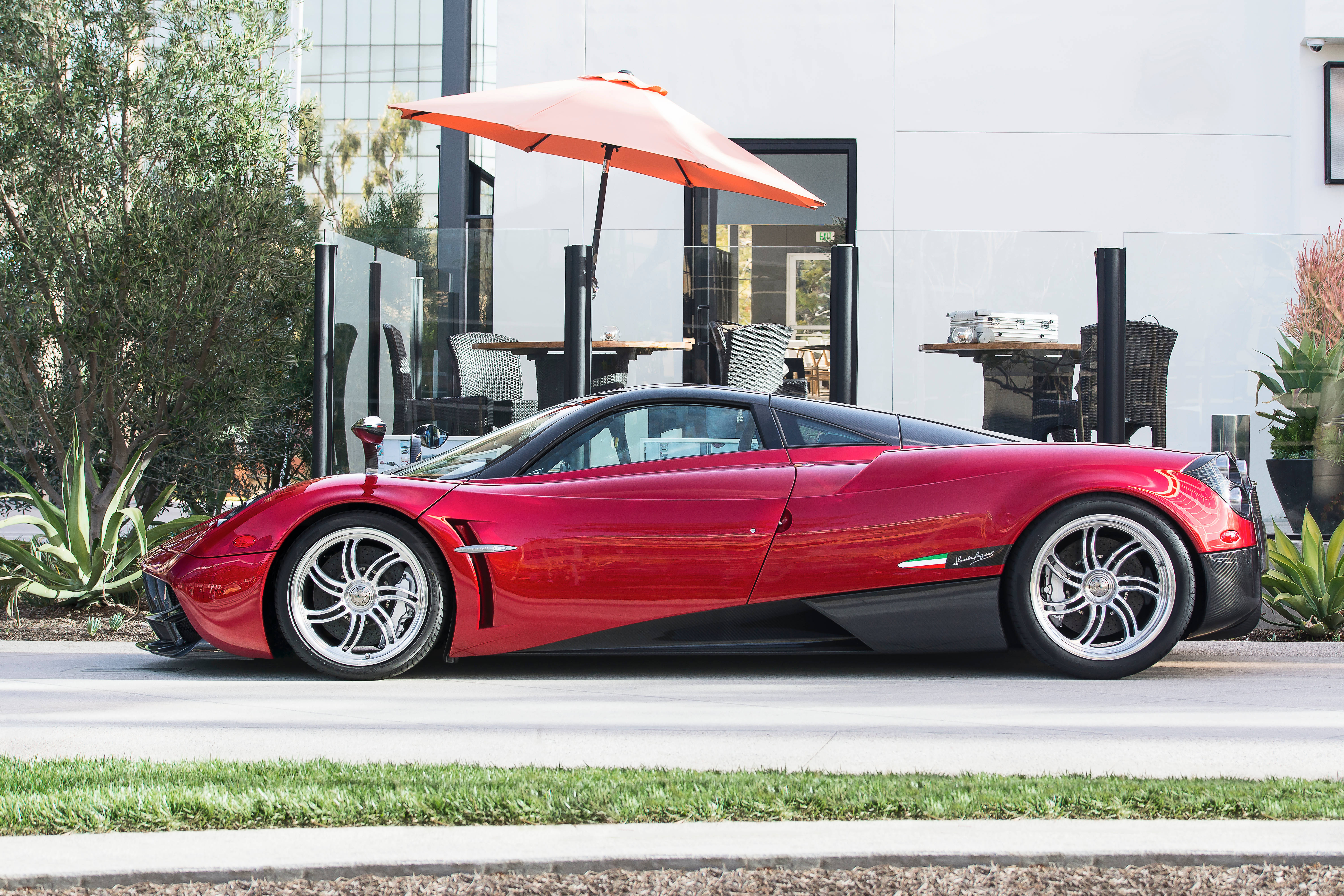 pagani iPhone 桌布 壁紙 — 一輛光澤的紅色Pagani Huayra跑車停在一棟現代建築旁邊，建築帶有橙色雨傘和綠色植物。