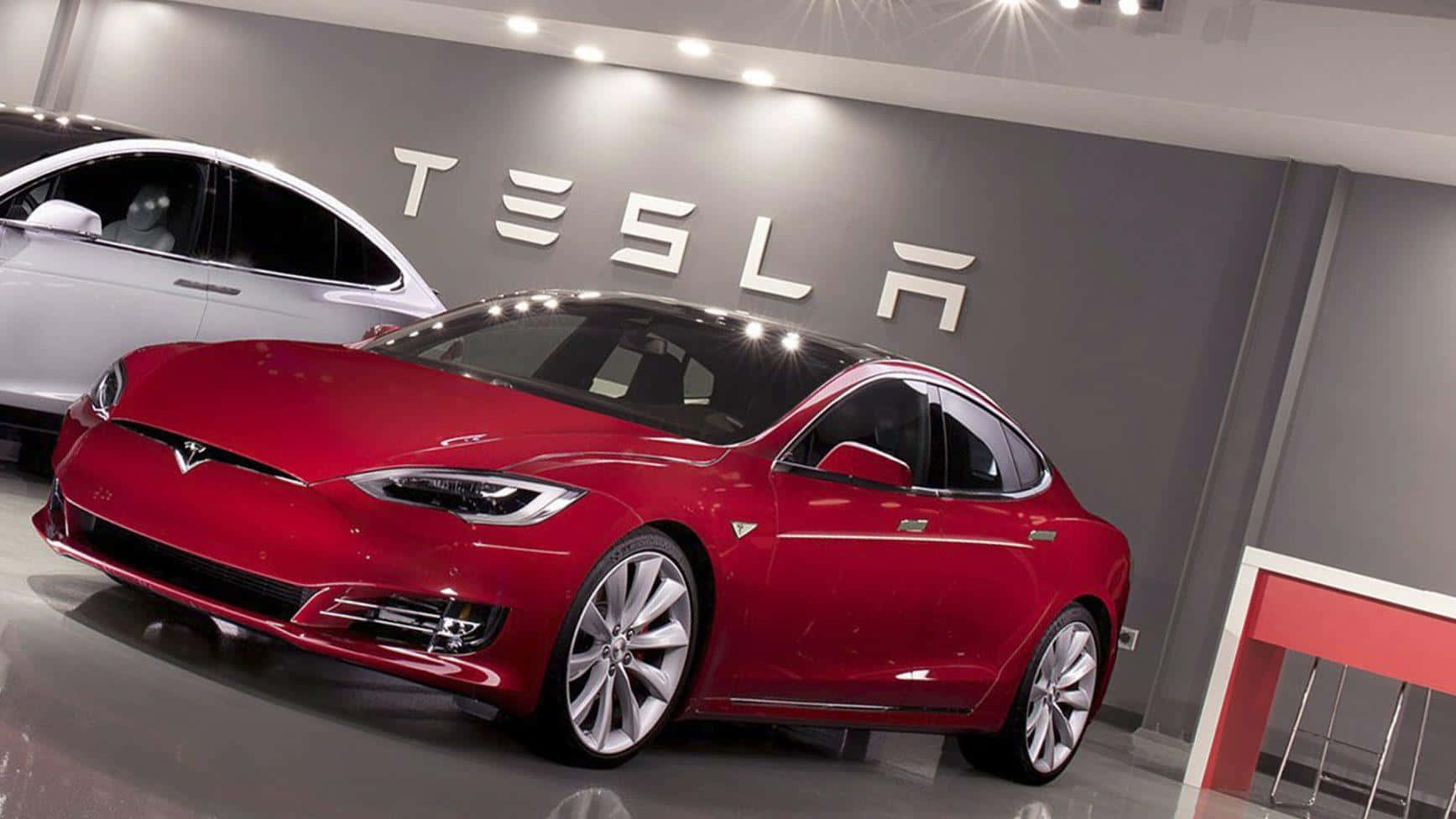 Tesla Model 3 桌布 壁紙 — 一輛光澤的紅色Tesla Model S轎車在展廳燈光下閃閃發光，後面的灰色牆上有白色的Tesla標誌。