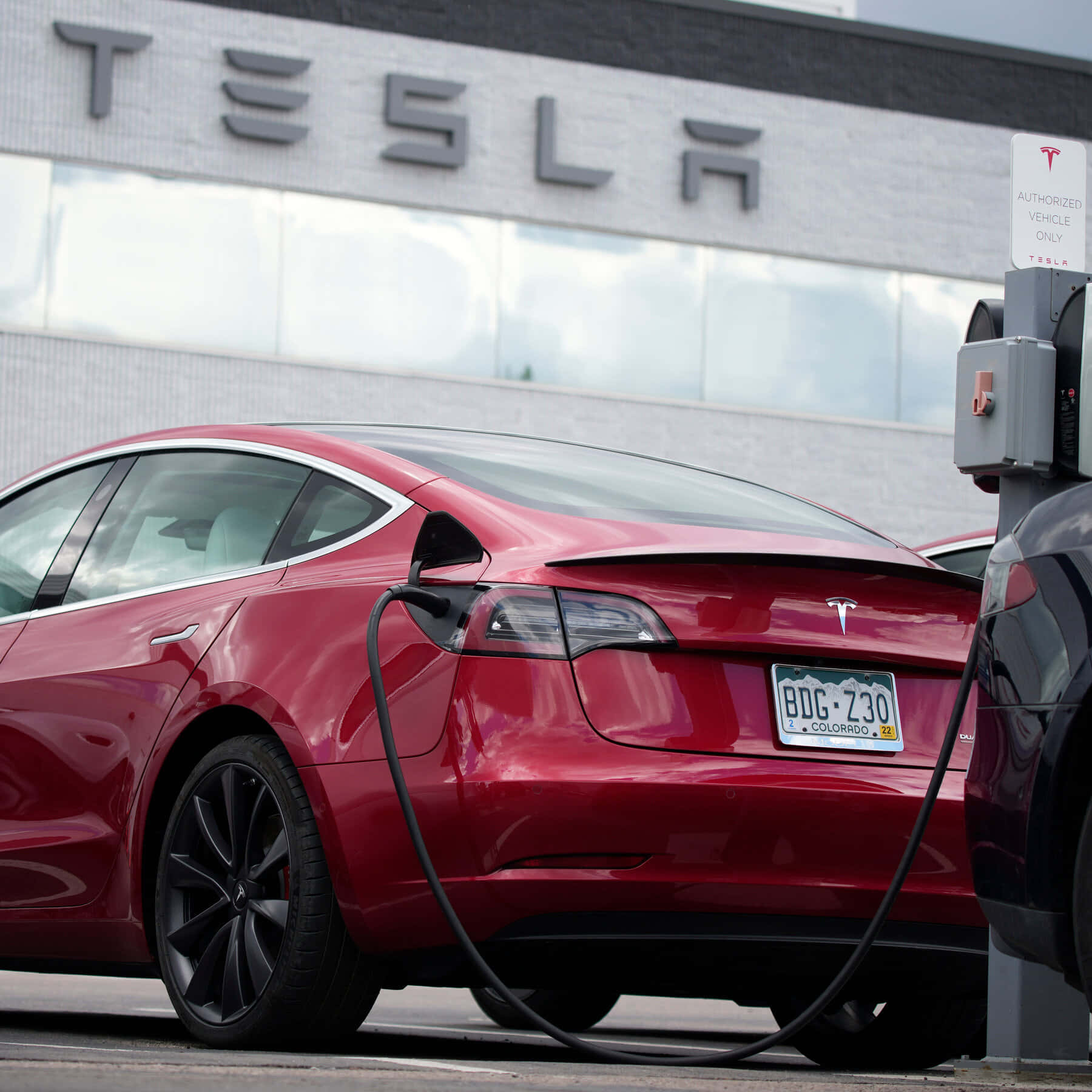 特斯拉 桌布 壁紙 — 一輛光澤的紅色Tesla Model S正在Tesla Supercharger站充電，後方建築物上可見品牌標誌。