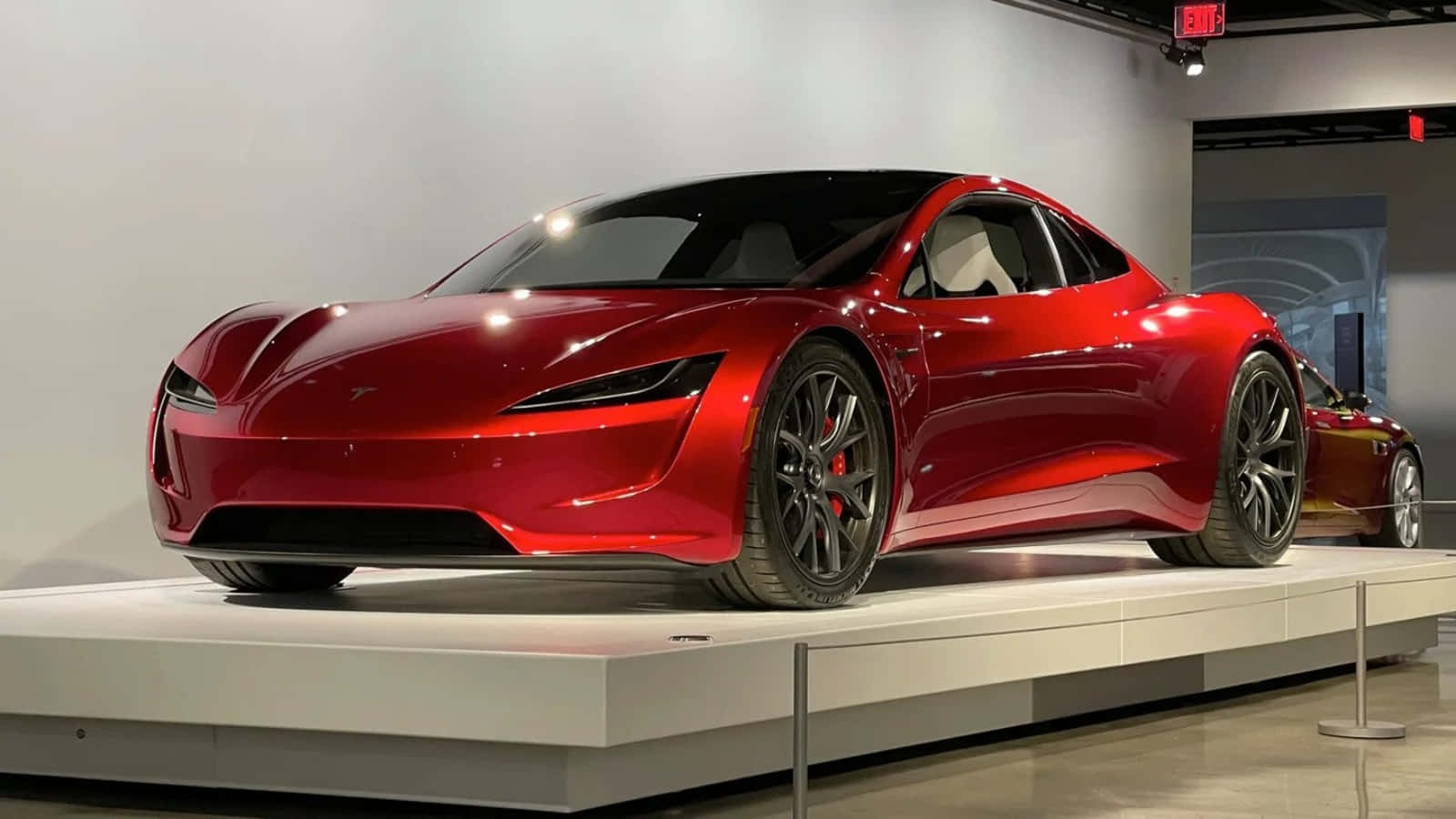 特斯拉 桌布 壁紙 — 一輛光澤的紅色Tesla Roadster跑車展示在現代展廳的白色平台上，光線明亮。